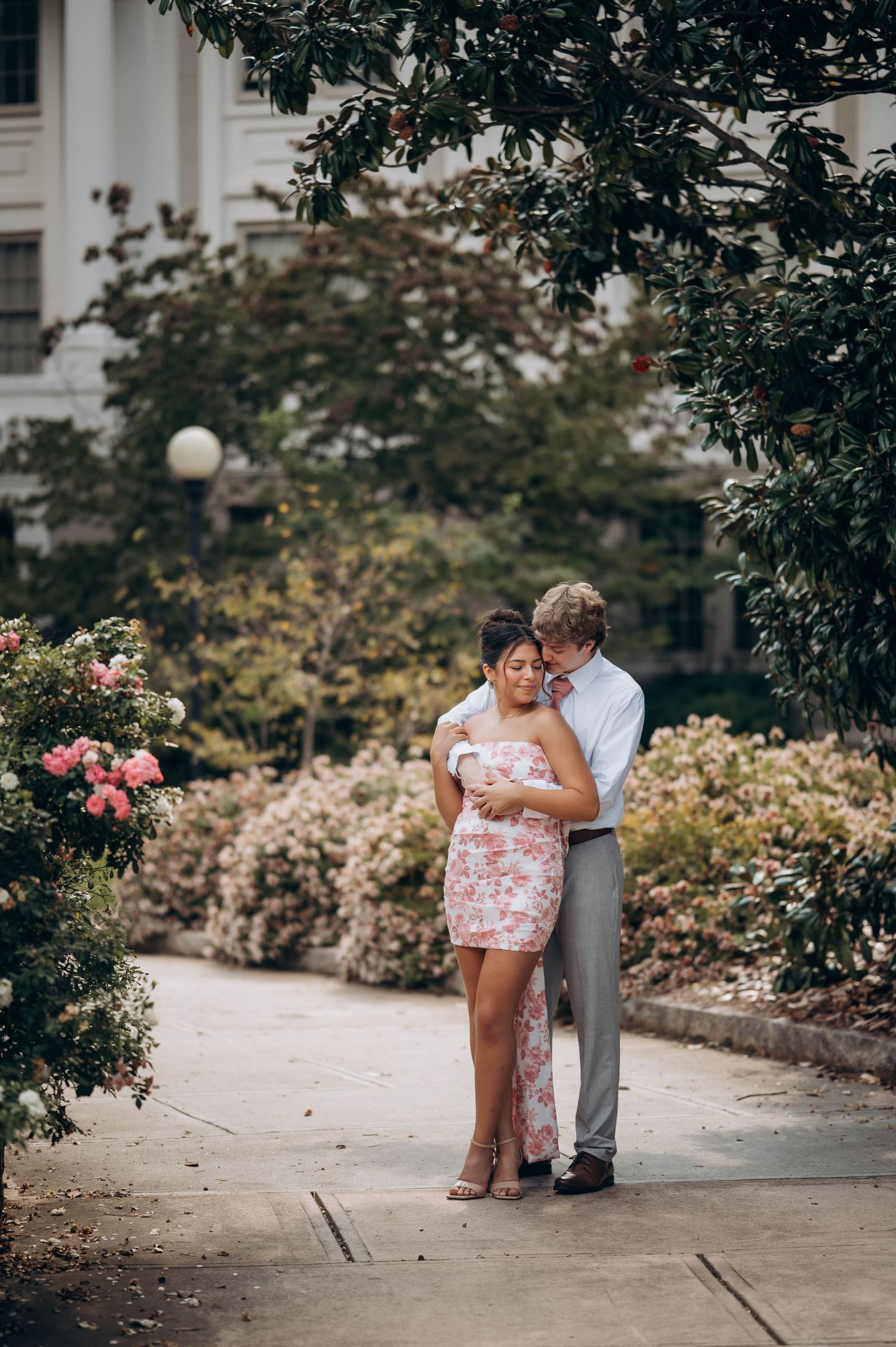 A romantic couple poses together on a pathway surrounded by lush garden landscaping.