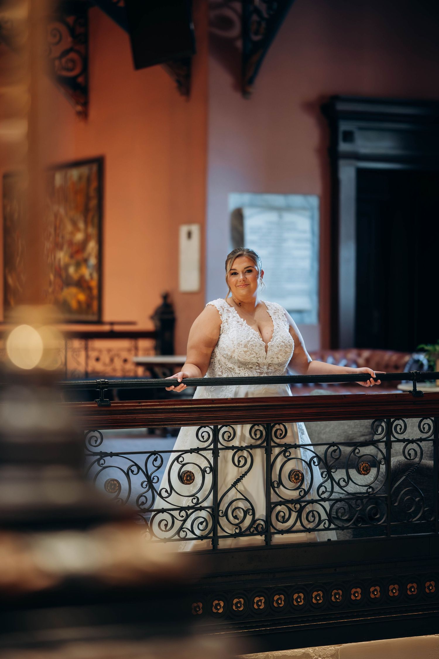 Bride in elegant white dress poses by decorative iron railing in warm-lit interior.