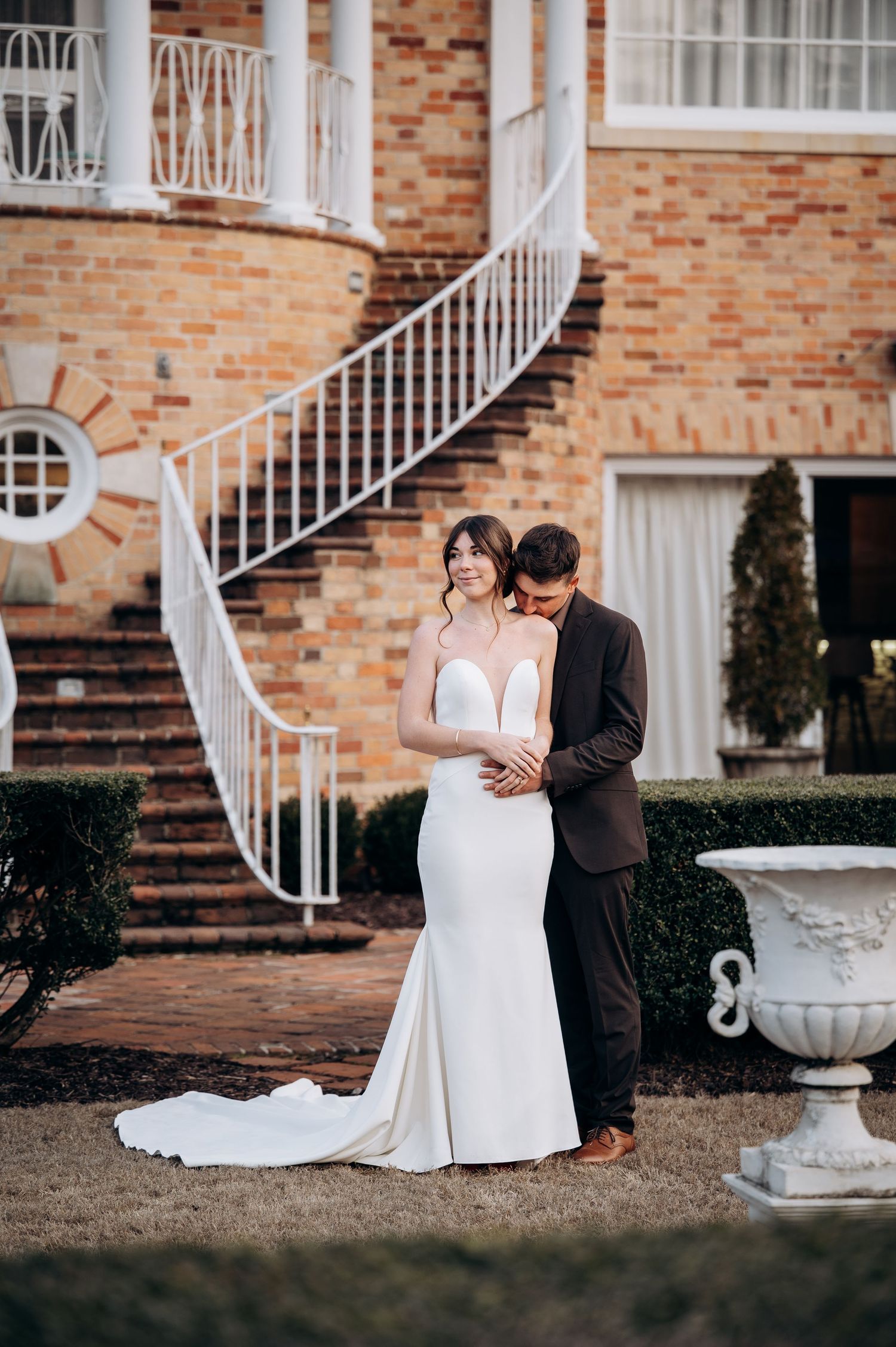 A bride in a white gown and groom in a dark suit embrace outside a brick mansion with a spiral staircase.