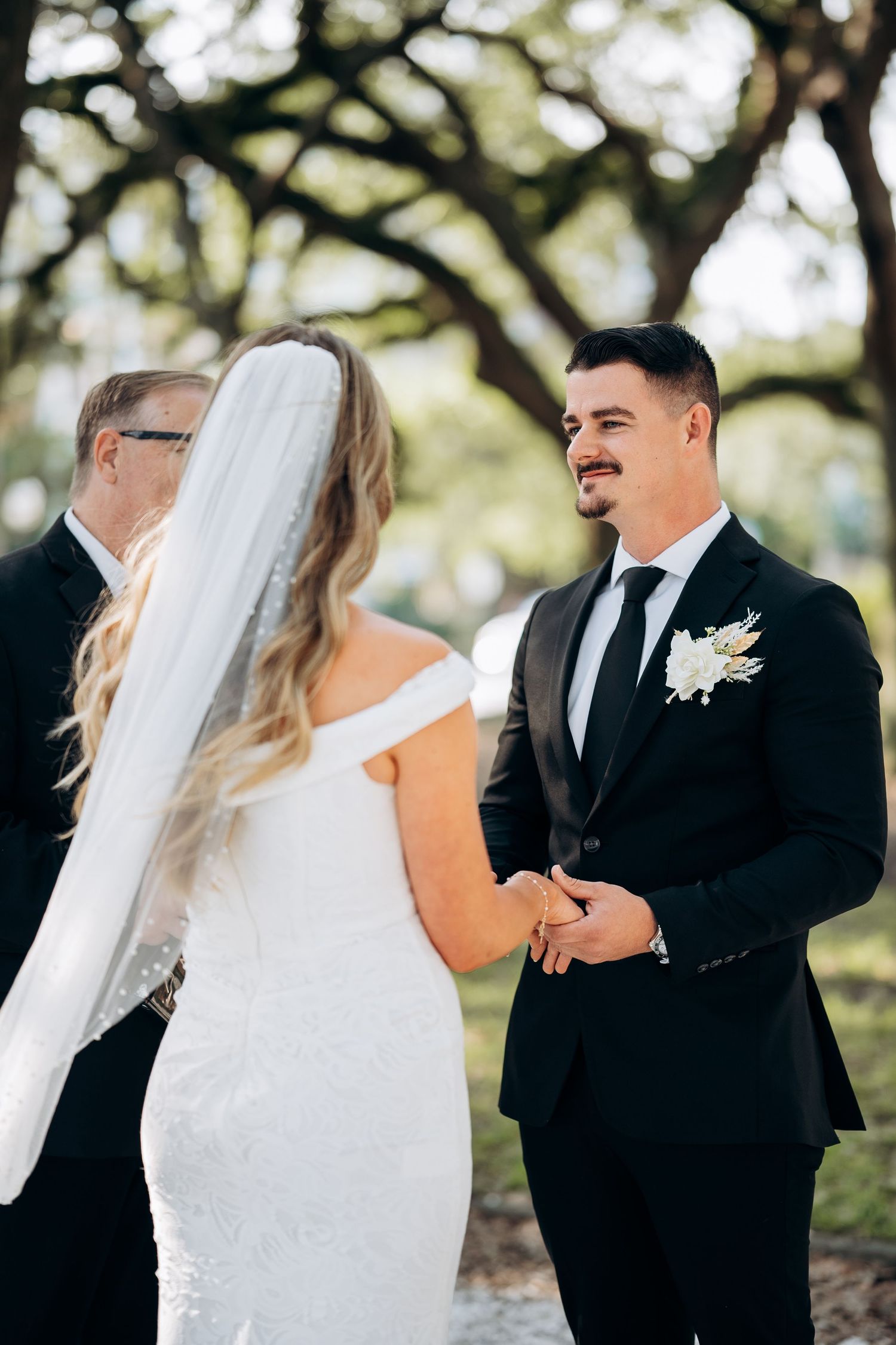 Smiling groom in black suit holds bride's hands at outdoor wedding ceremony under oak trees.