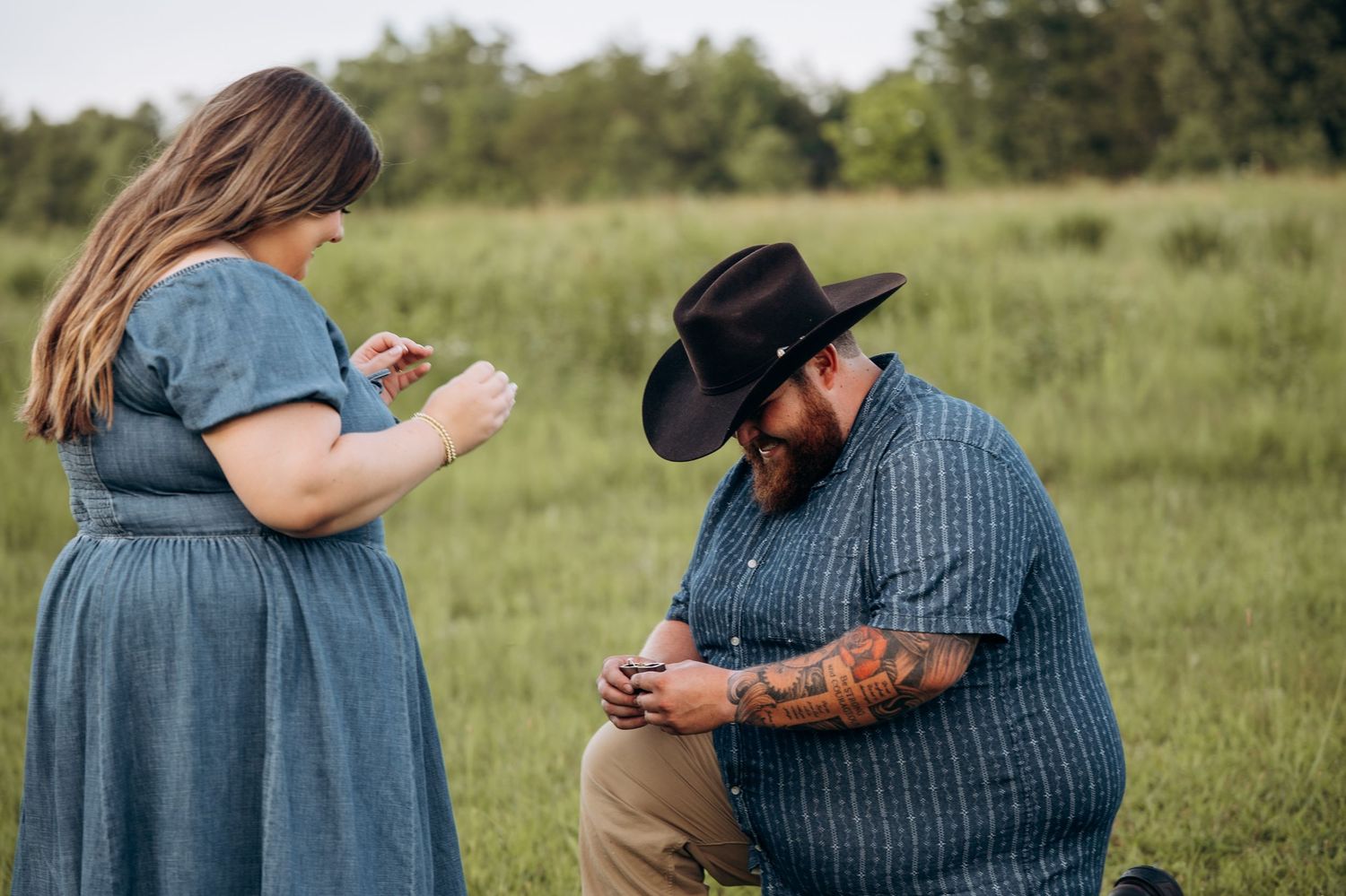 Man in cowboy hat kneeling to propose to woman in blue dress in a green field outdoor engagement photo.