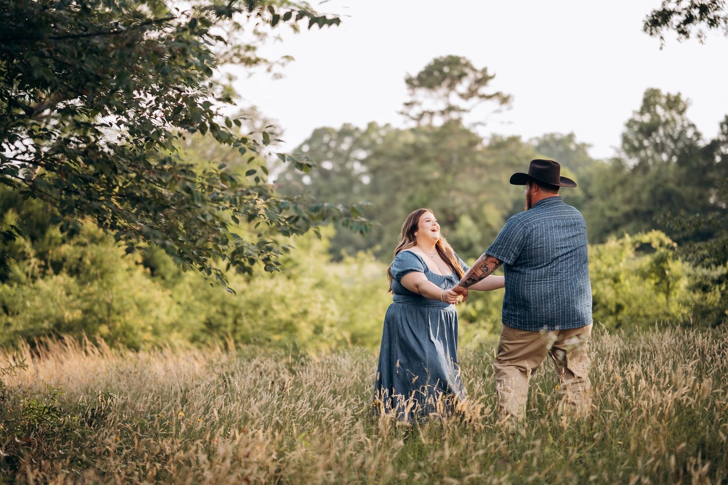 Couple holding hands and dancing in a golden grass field surrounded by lush green trees.