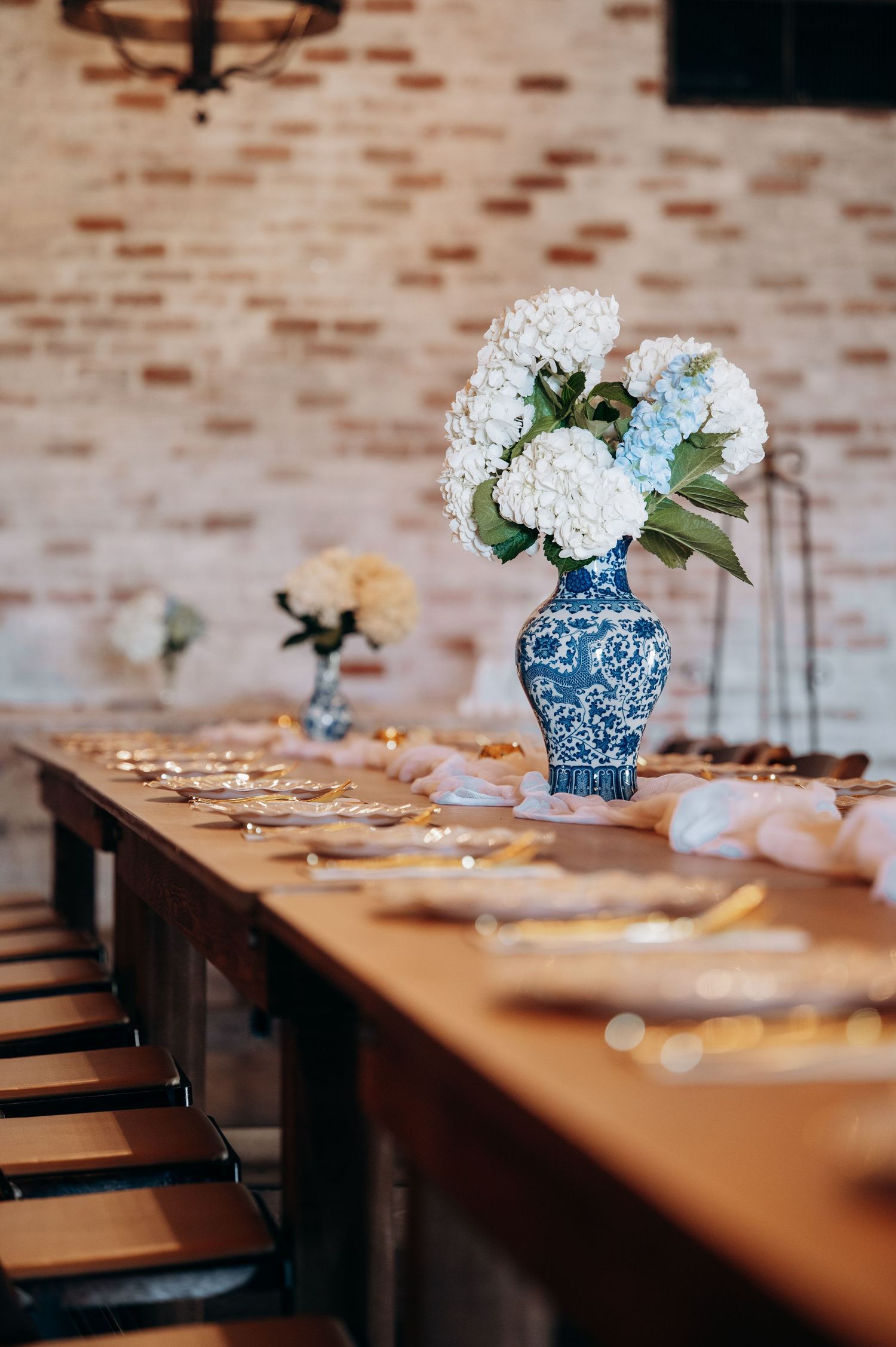 Blue and white chinoiserie vase with white hydrangeas centerpiece on wooden table with gold accents and brick wall backdrop.