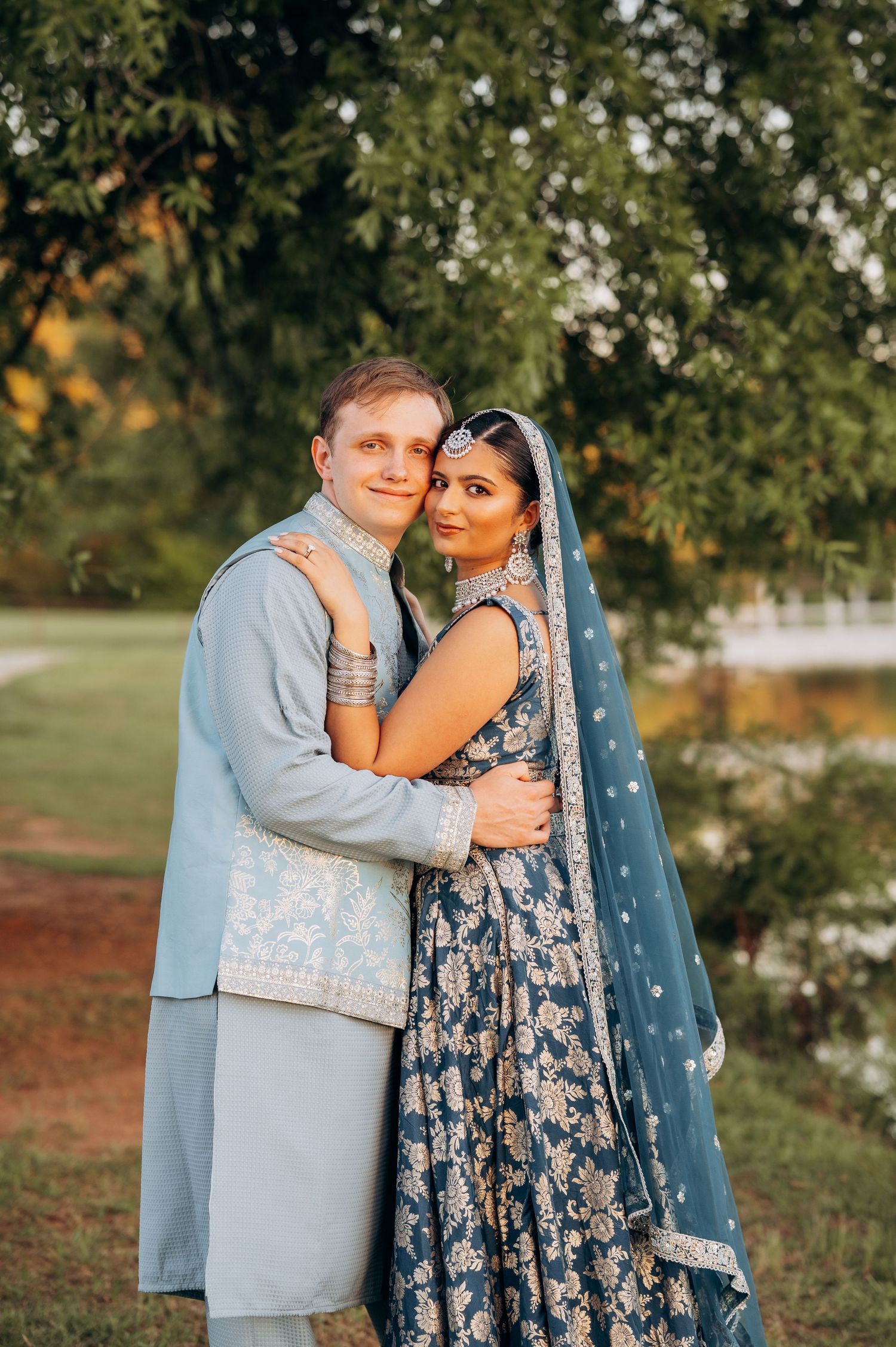 Couple in traditional South Asian wedding attire poses outdoors, man in light blue sherwani, woman in navy lehenga.