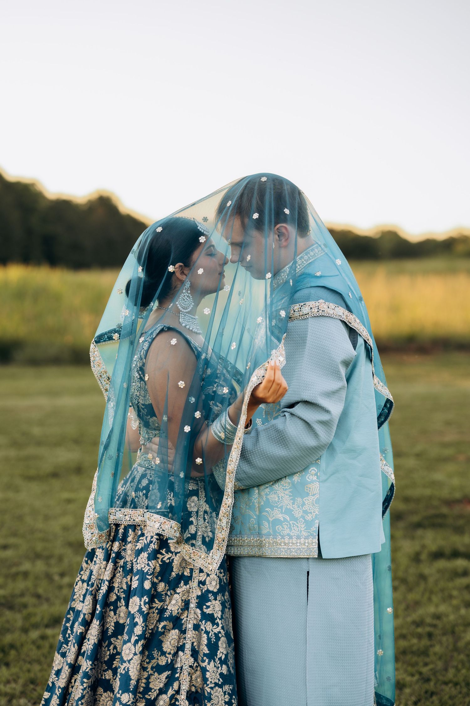 Indian bride and groom share romantic moment under blue veil in traditional wedding attire outdoors.