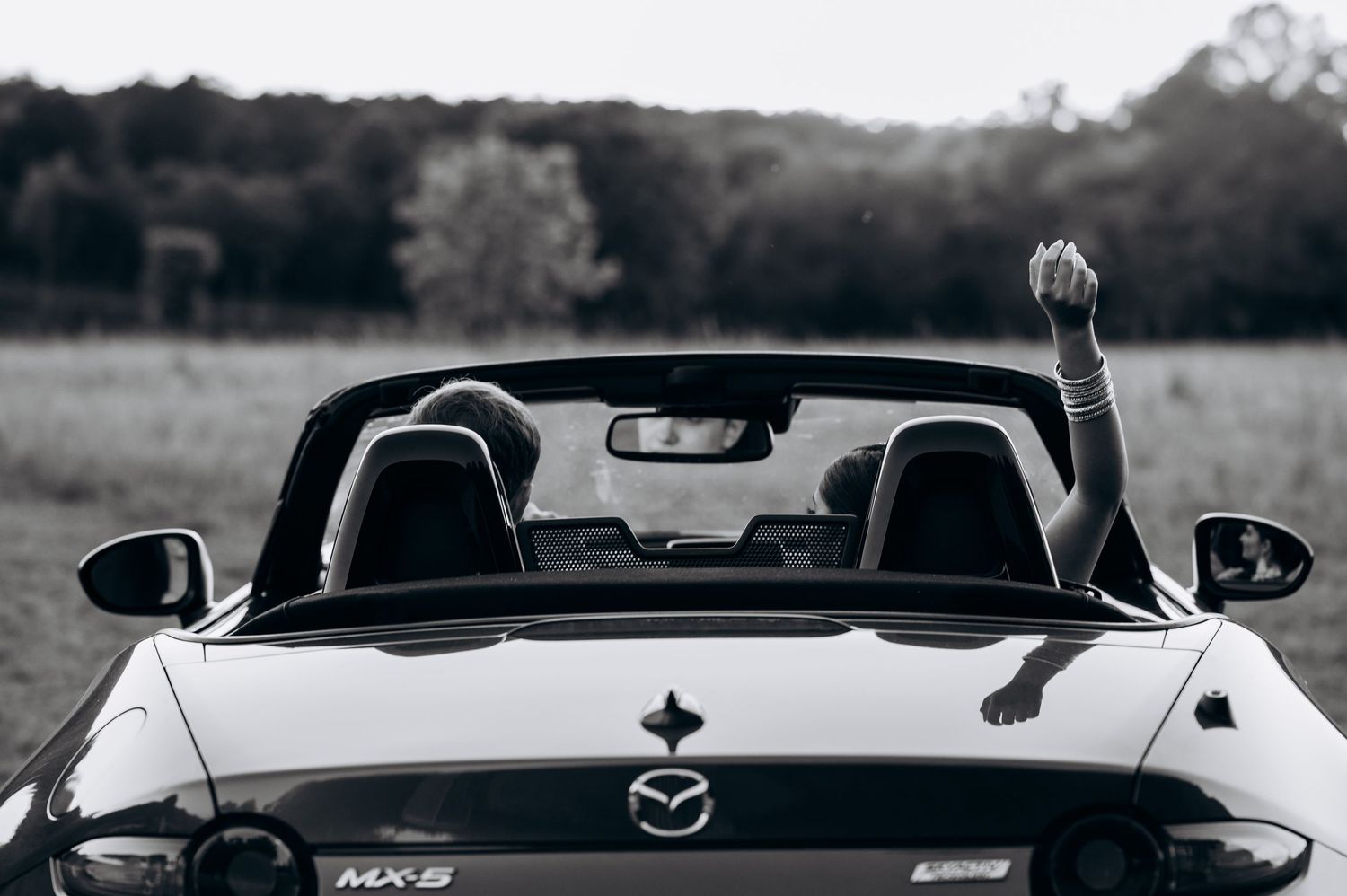 Black and white photo of a Mazda MX-5 convertible with a passenger raising their hand in an open field.
