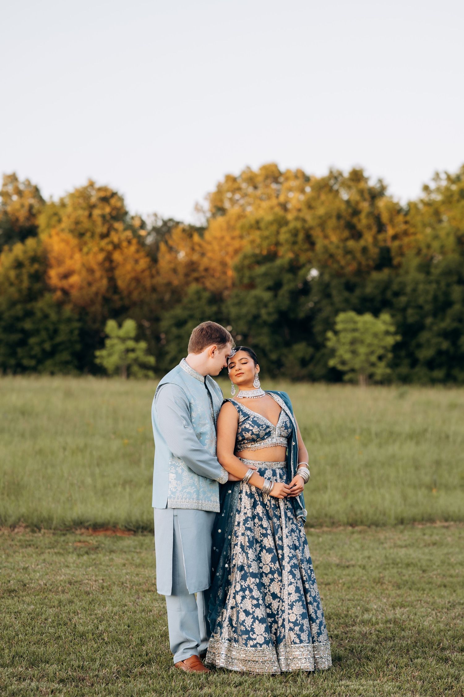 Couple in blue Indian wedding attire embracing in a green field at golden hour, with trees in the background.