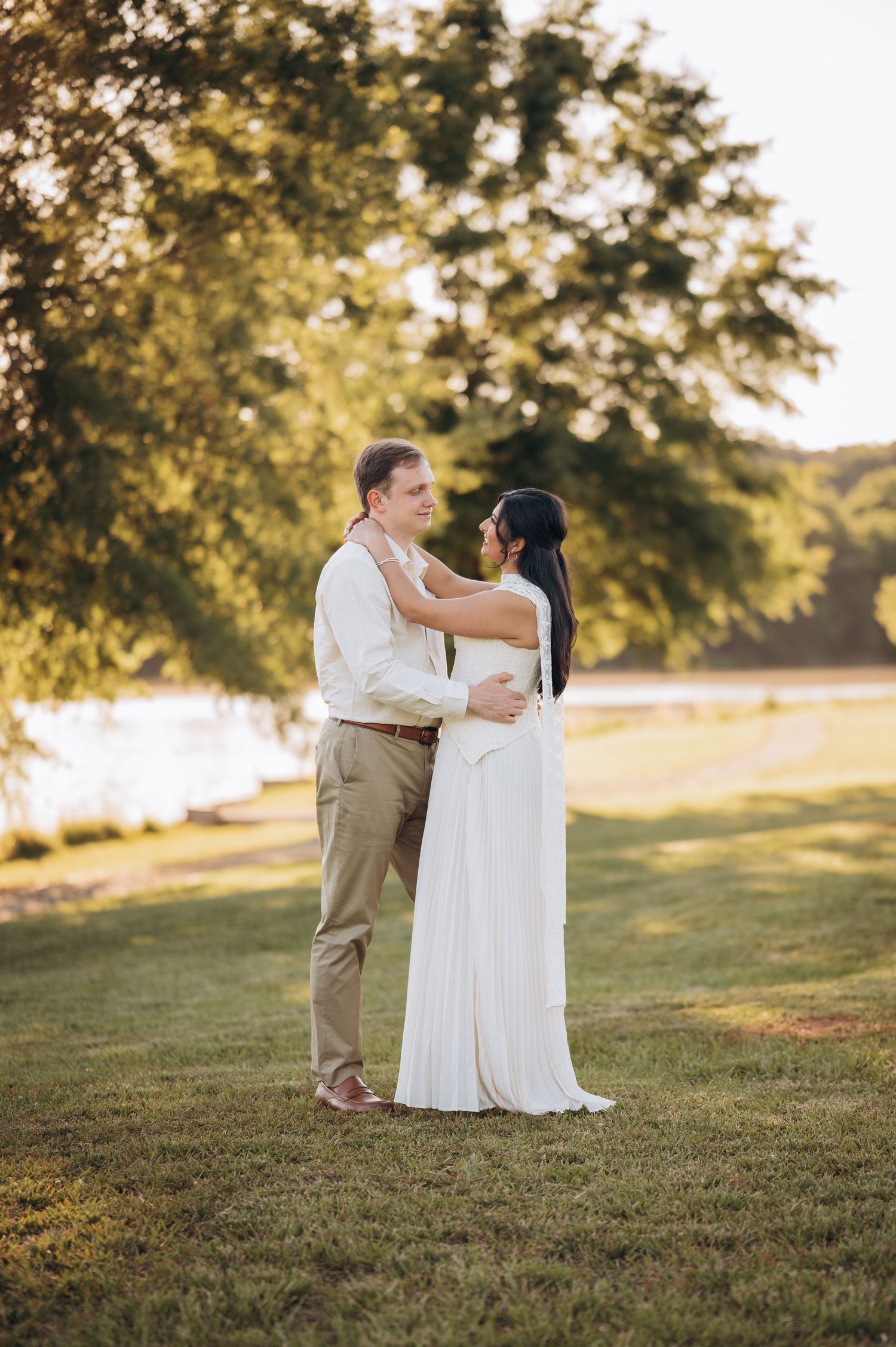 Couple embracing in white wedding attire on green lawn with golden sunset light filtering through lush trees near water.