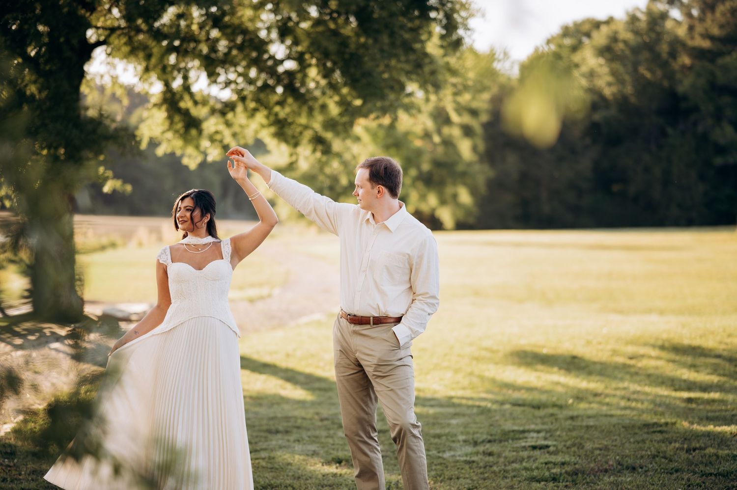 Bride and groom dancing outdoors in lush green park, groom spinning bride in white pleated wedding dress on sunny day.