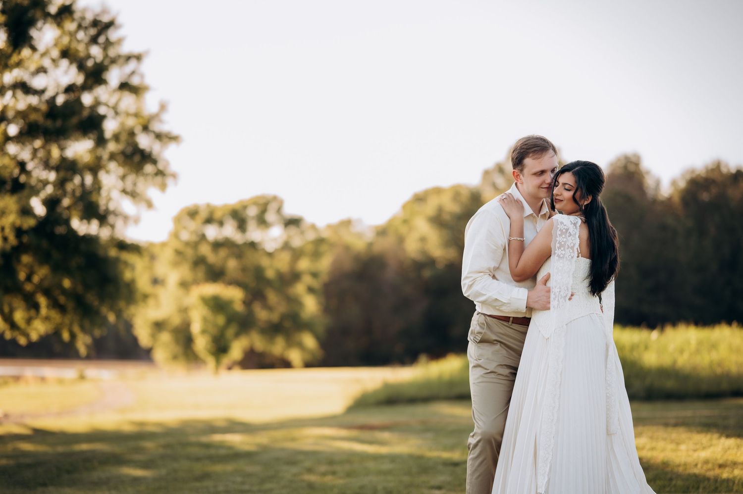 Bride and groom share a romantic embrace outdoors at golden hour, surrounded by lush green trees and meadow.