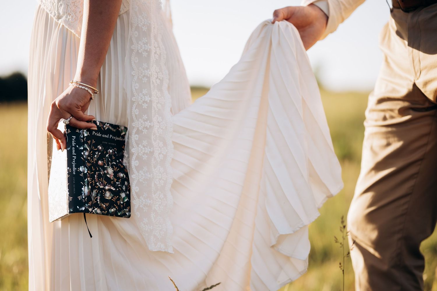 Bride in white lace gown holding clutch purse with groom lifting dress hem in sunny field.