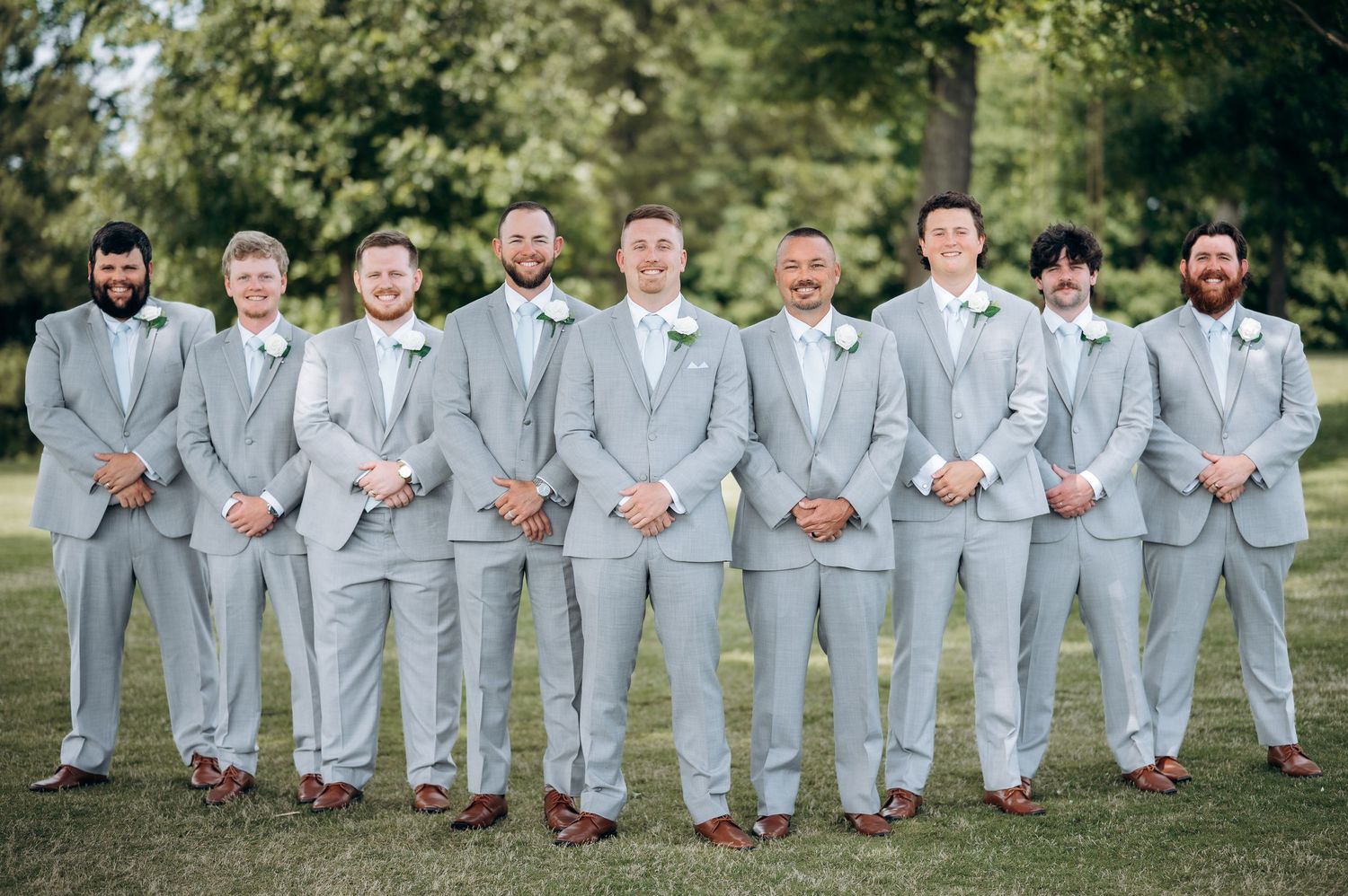 Group of groomsmen posing outdoors in matching light gray suits with white boutonnieres on green lawn.