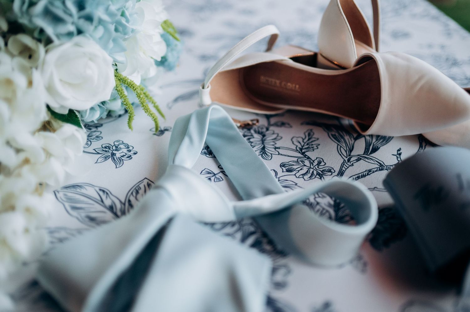 Elegant bridal accessories including nude heels, blue floral ribbon, and white bouquet arranged on patterned fabric.