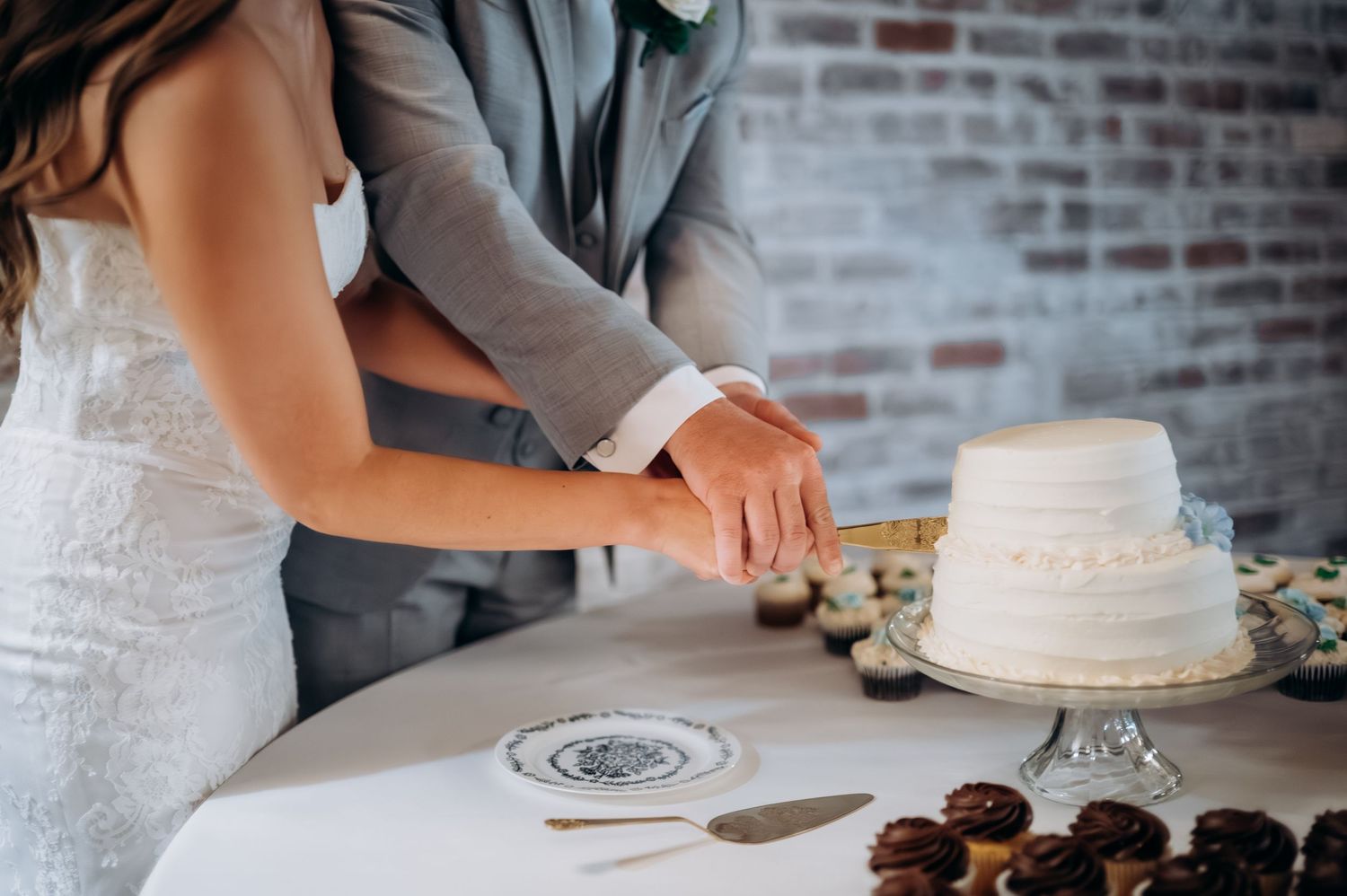 Bride and groom cutting white two-tier wedding cake together at reception table with cupcakes nearby.