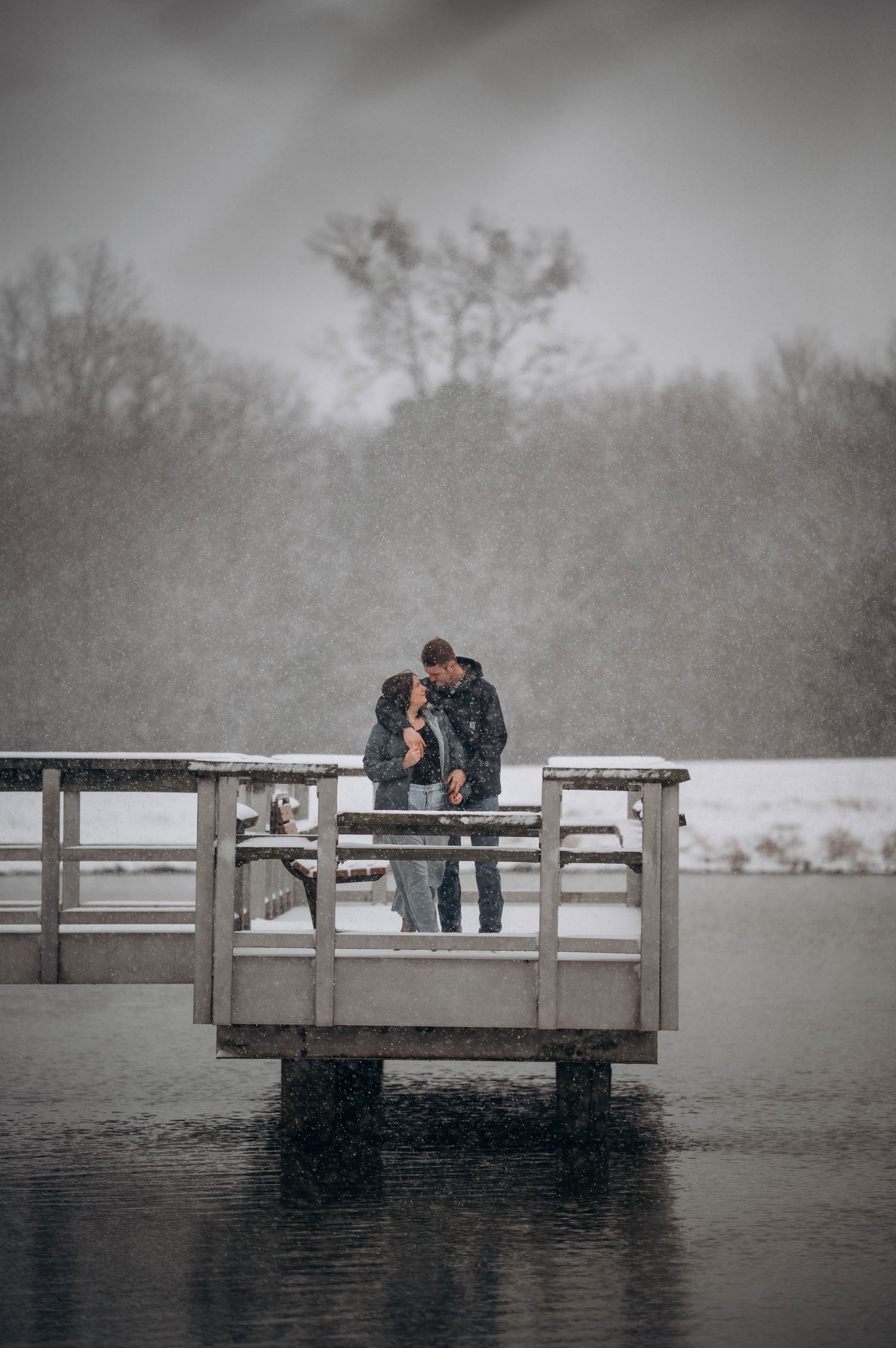 A couple embraces on a wooden dock over a misty lake during winter, creating a romantic snowy atmosphere.