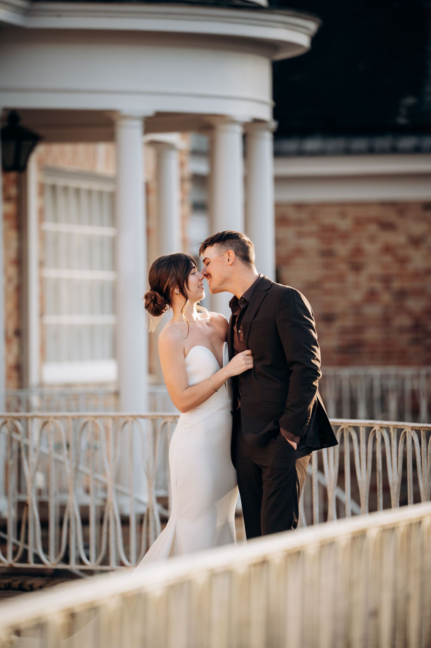 A romantic wedding couple shares a tender kiss on an elegant columned porch with white railings.