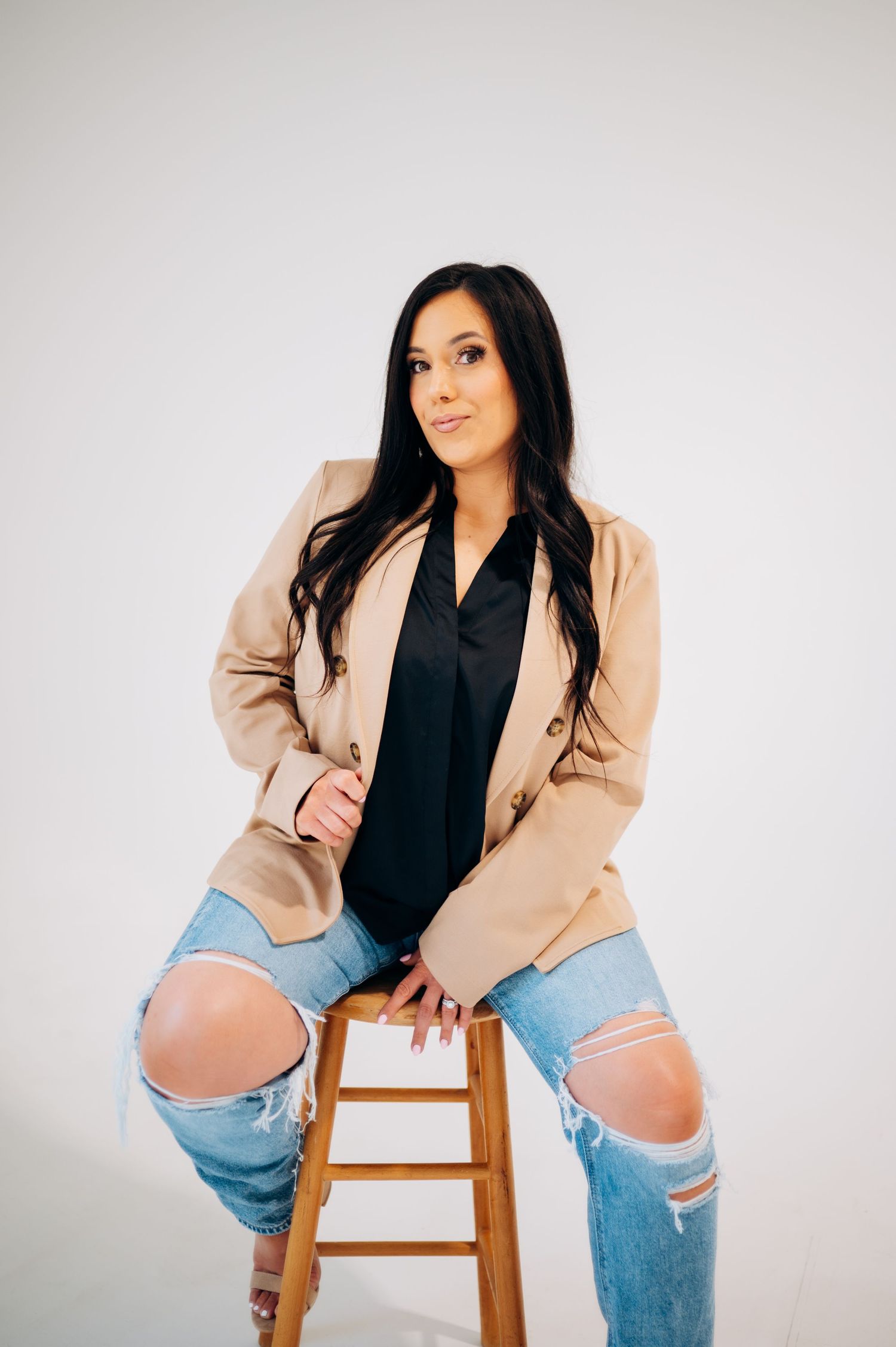 Stylish outfit featuring tan blazer, black top and ripped jeans while seated on wooden stool.