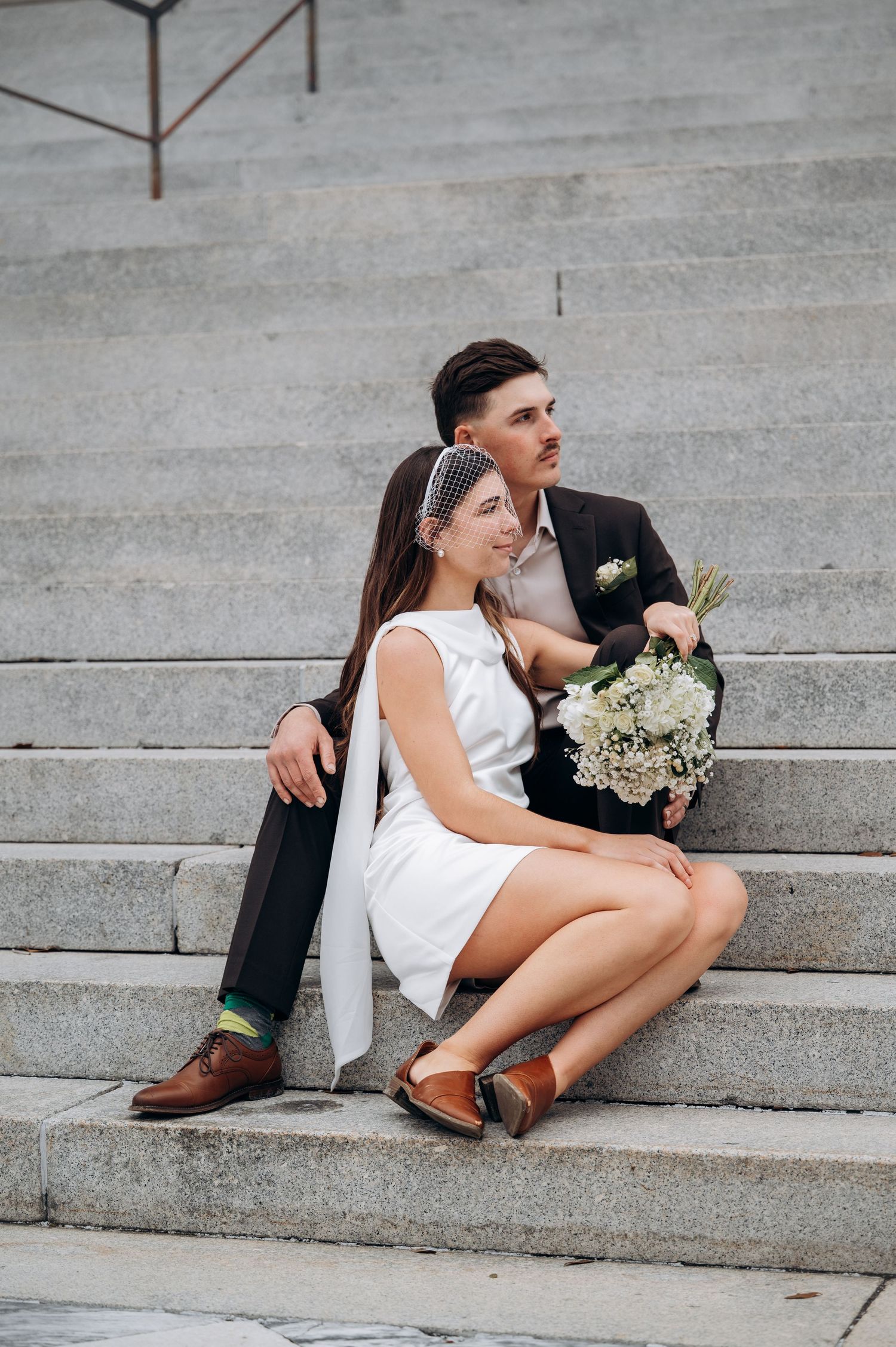 Couple in wedding attire sitting casually on concrete steps during photo session.
