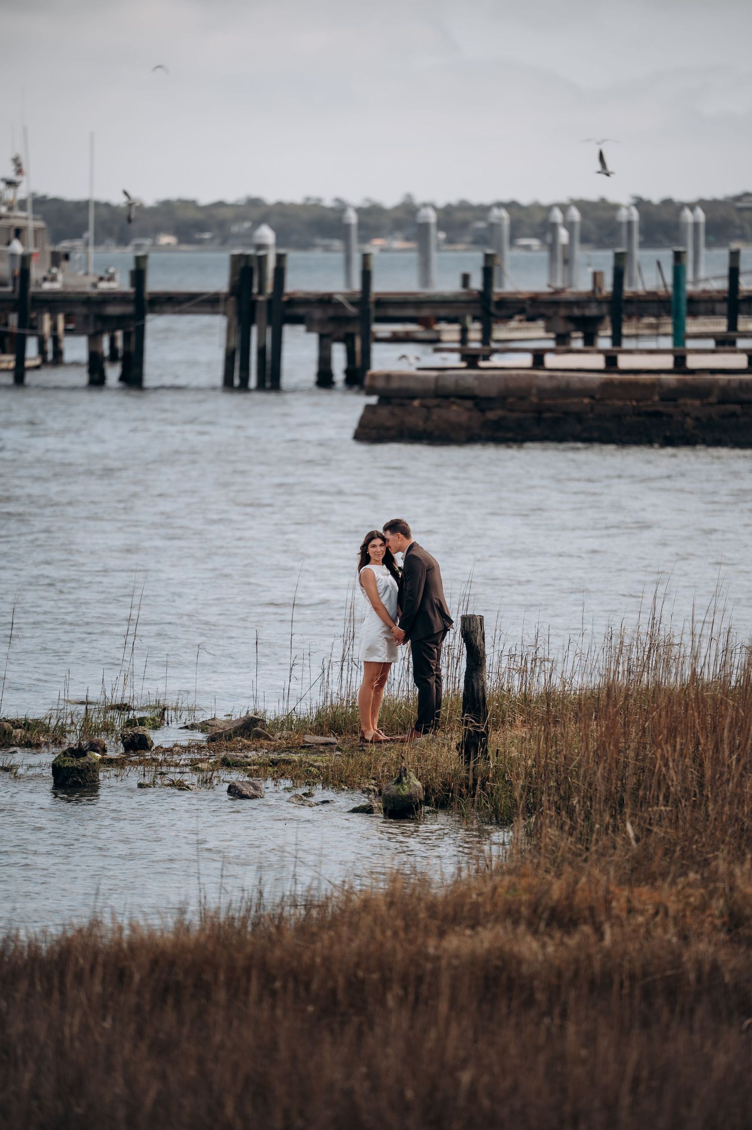 A couple shares an intimate moment on a wooden pier by the water with tall marsh grass in the foreground.