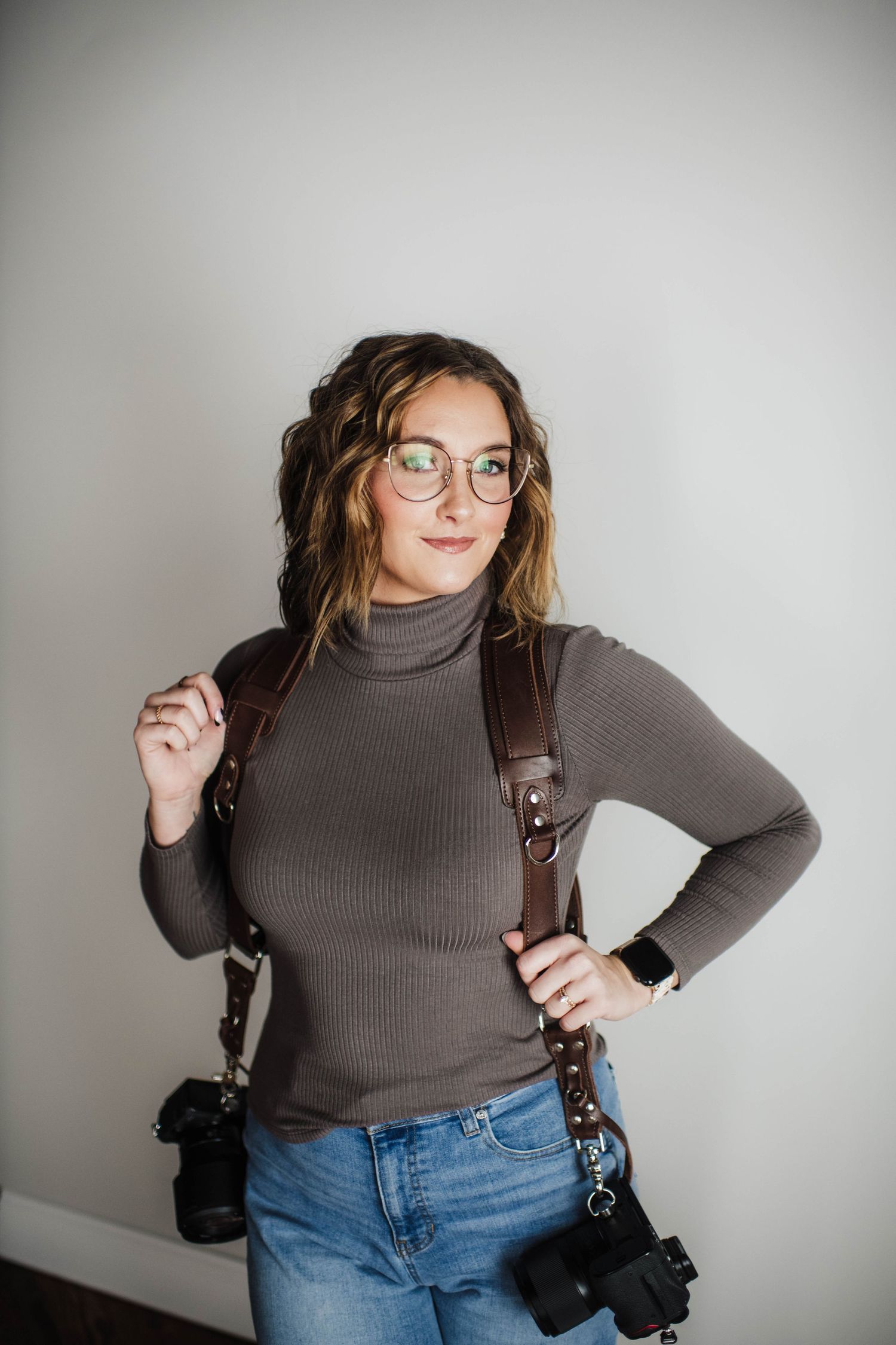 Woman photographer wearing glasses and turtleneck with dual camera harness and DSLR cameras, posing confidently.