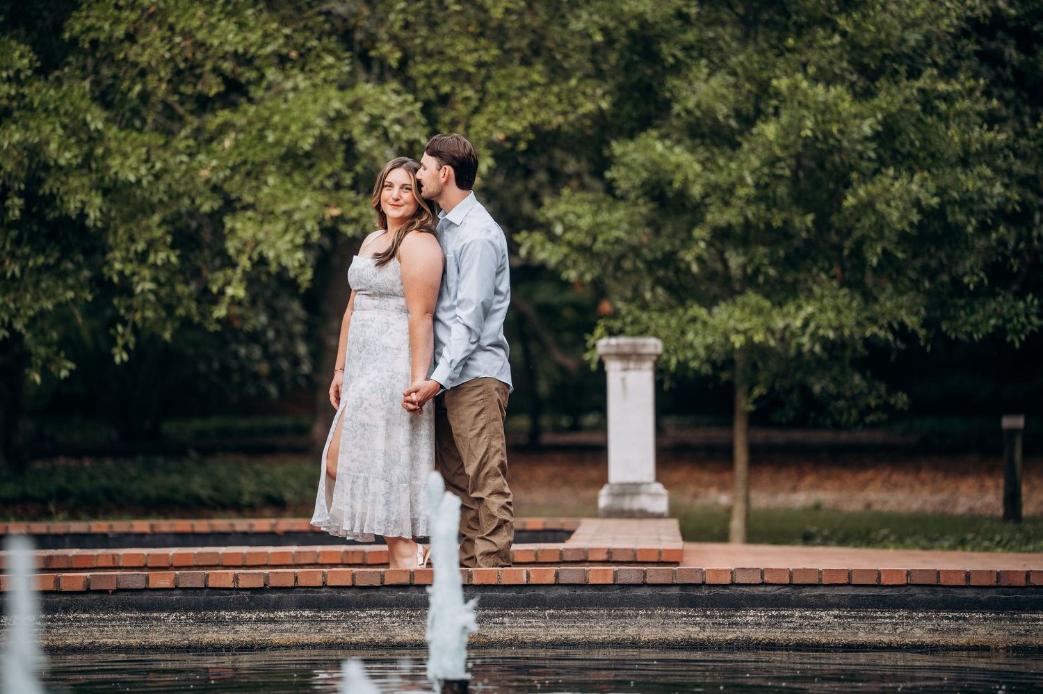 Engaged couple walking hand in hand near fountain in lush green park setting.