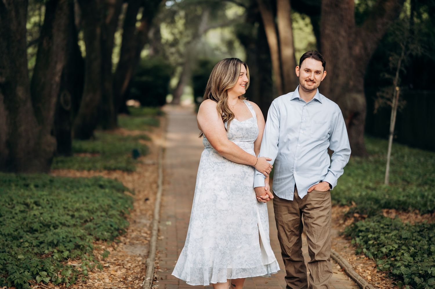 Couple holding hands on a tree-lined path, woman in white dress and man in blue shirt, outdoor engagement photo.