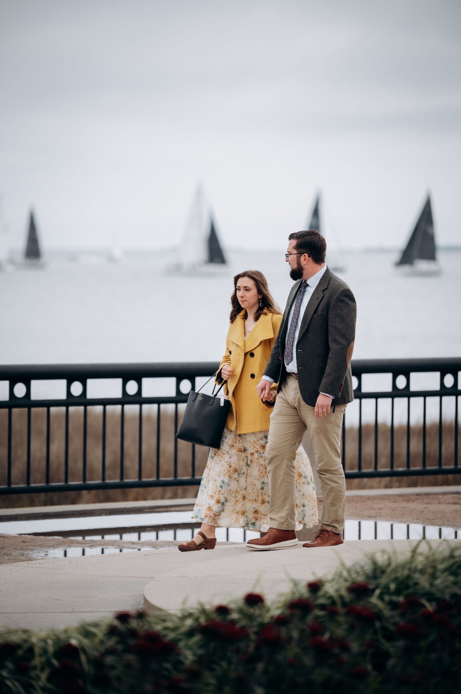 Couple walks along waterfront promenade, woman in yellow coat and floral skirt, sailboats behind.