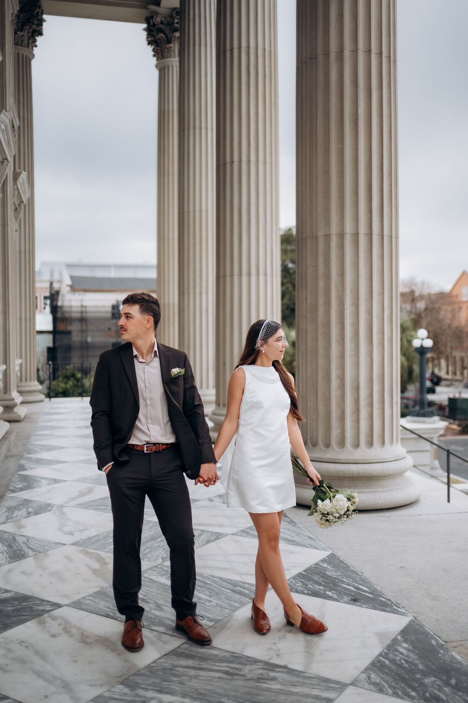 Bride and groom hold hands among grand columns on marble steps, she in a white mini dress holding a bouquet.
