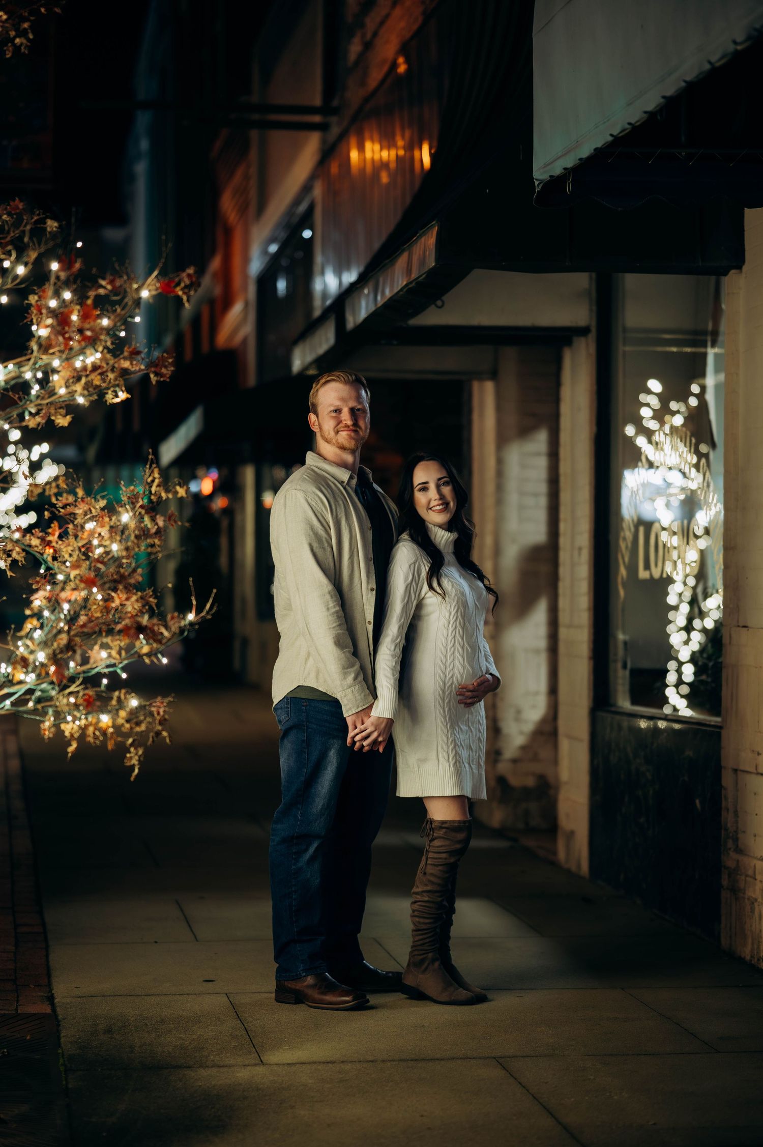 Couple holding hands on a festive night street, woman in white sweater dress and tall boots showing baby bump.
