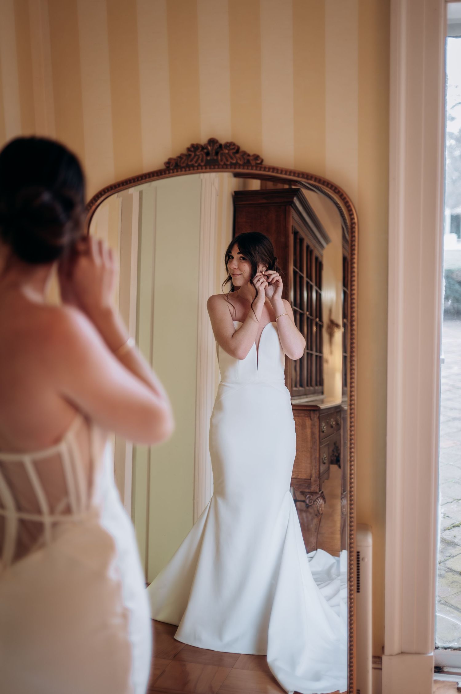 Bride in fitted white mermaid gown adjusts earring while viewing reflection in ornate gold floor mirror.