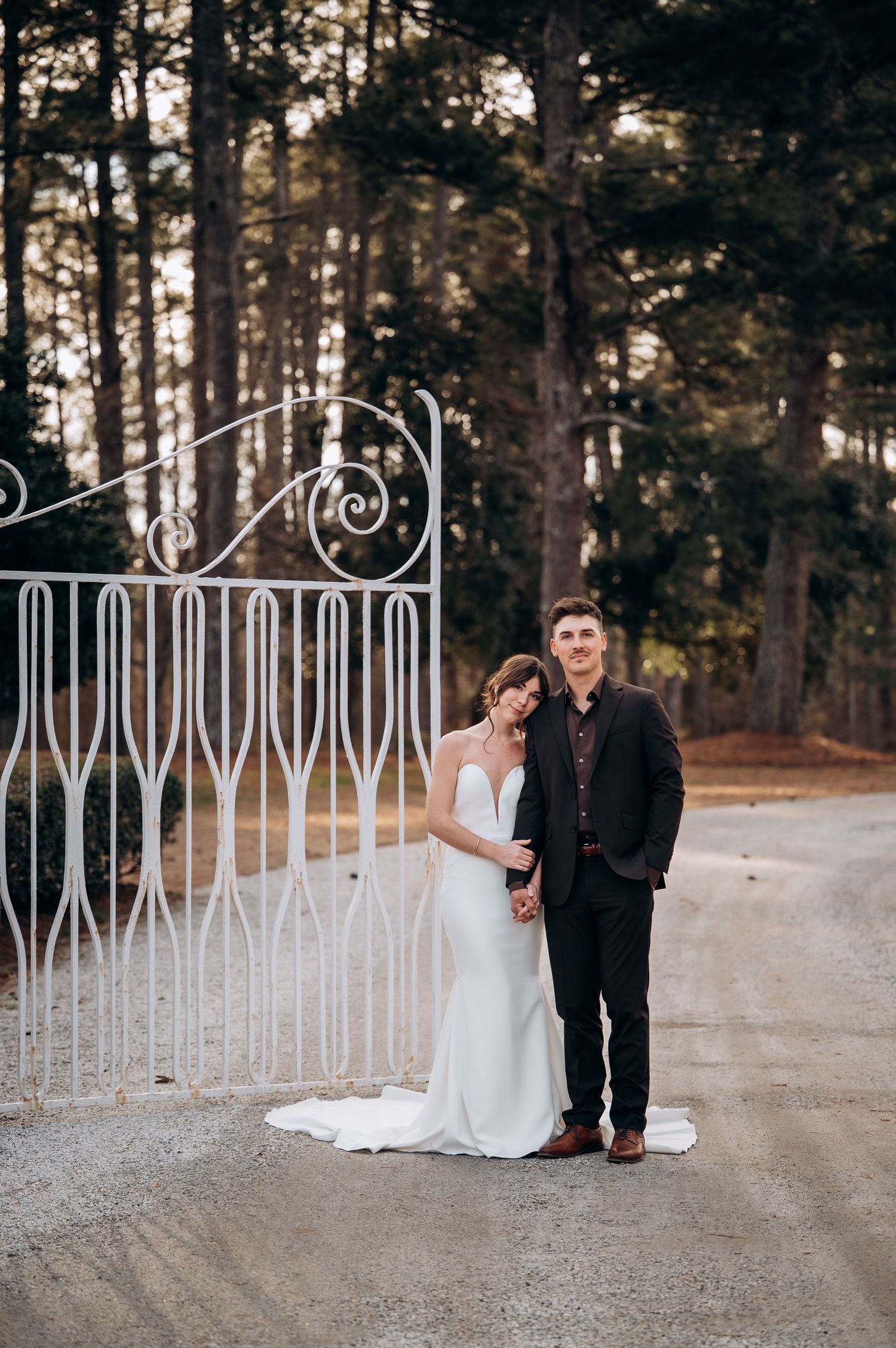 Bride and groom posing together before ornate white iron gate surrounded by tall pine trees.