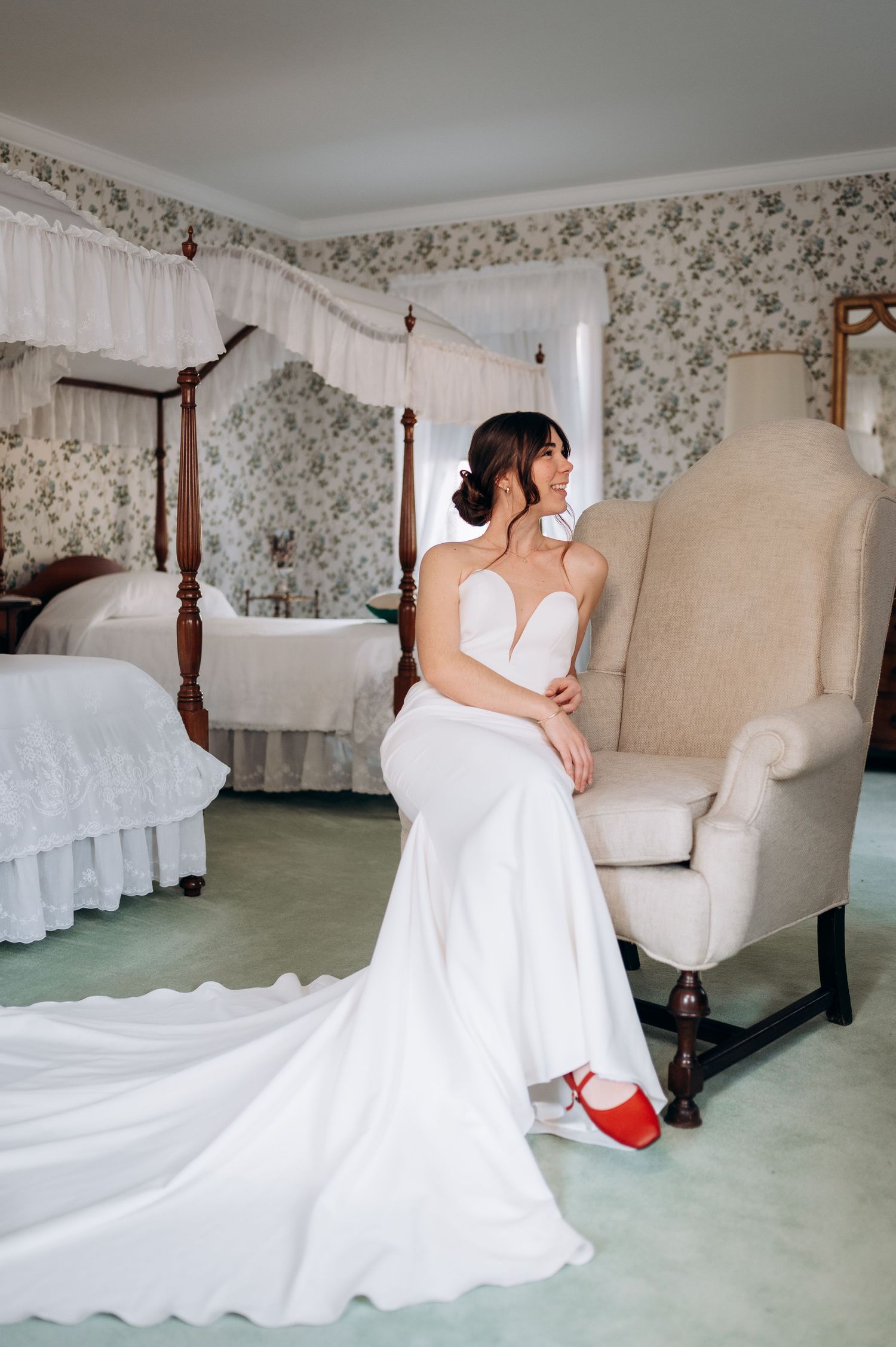 Bride in fitted white strapless gown with red shoes seated in wingback chair in floral wallpapered bedroom.