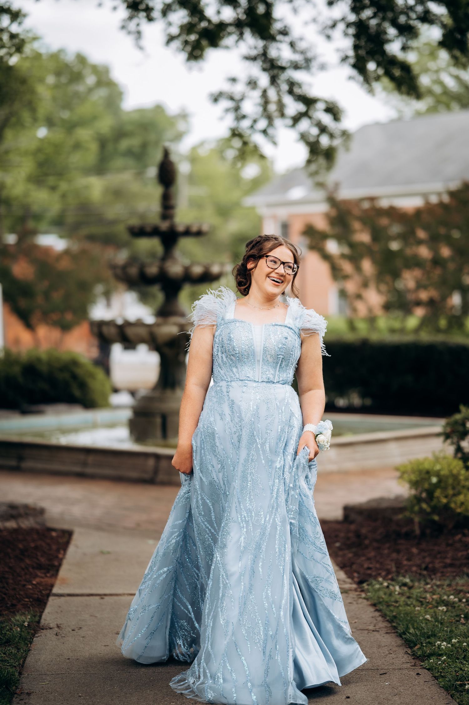 Young woman in light blue ball gown with feather details smiling in garden with fountain backdrop.