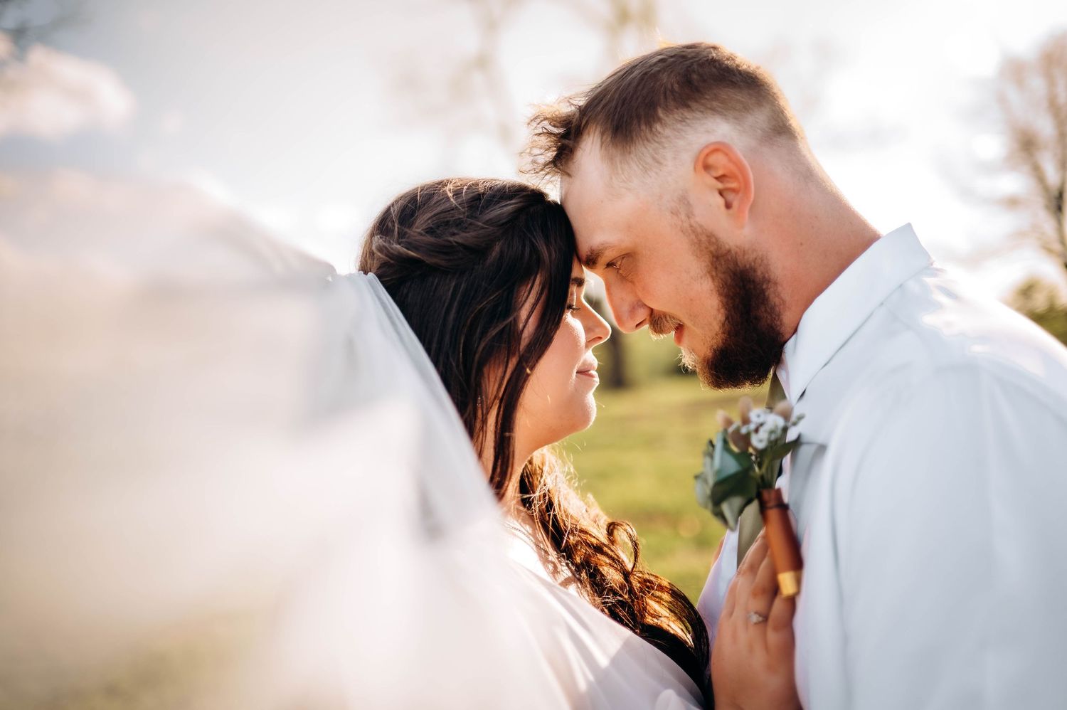 Romantic wedding couple touching foreheads outdoors, groom in white shirt with boutonniere, bride in veil, soft natural light.