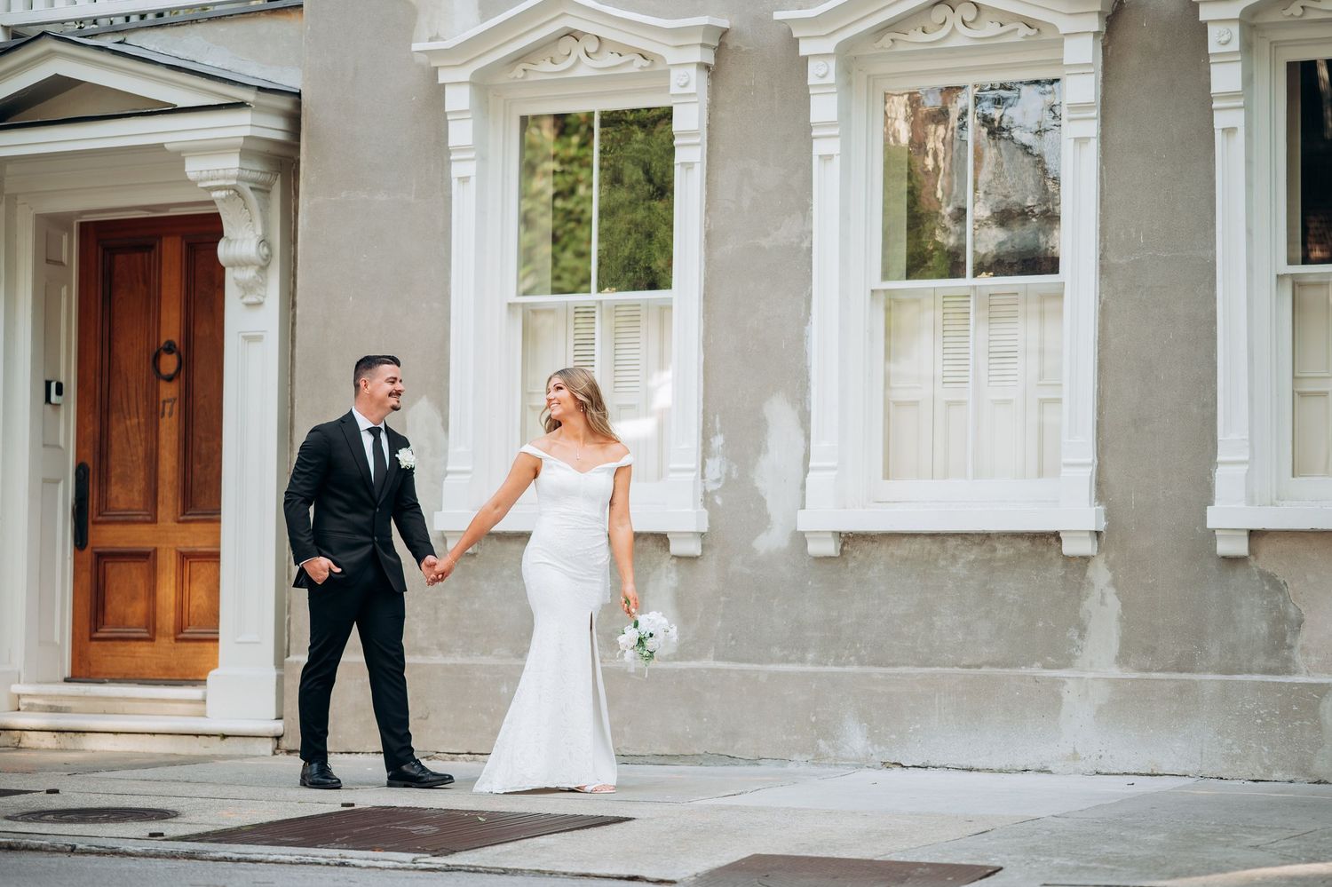 Bride and groom holding hands walking outside a classic grey building on their wedding day.
