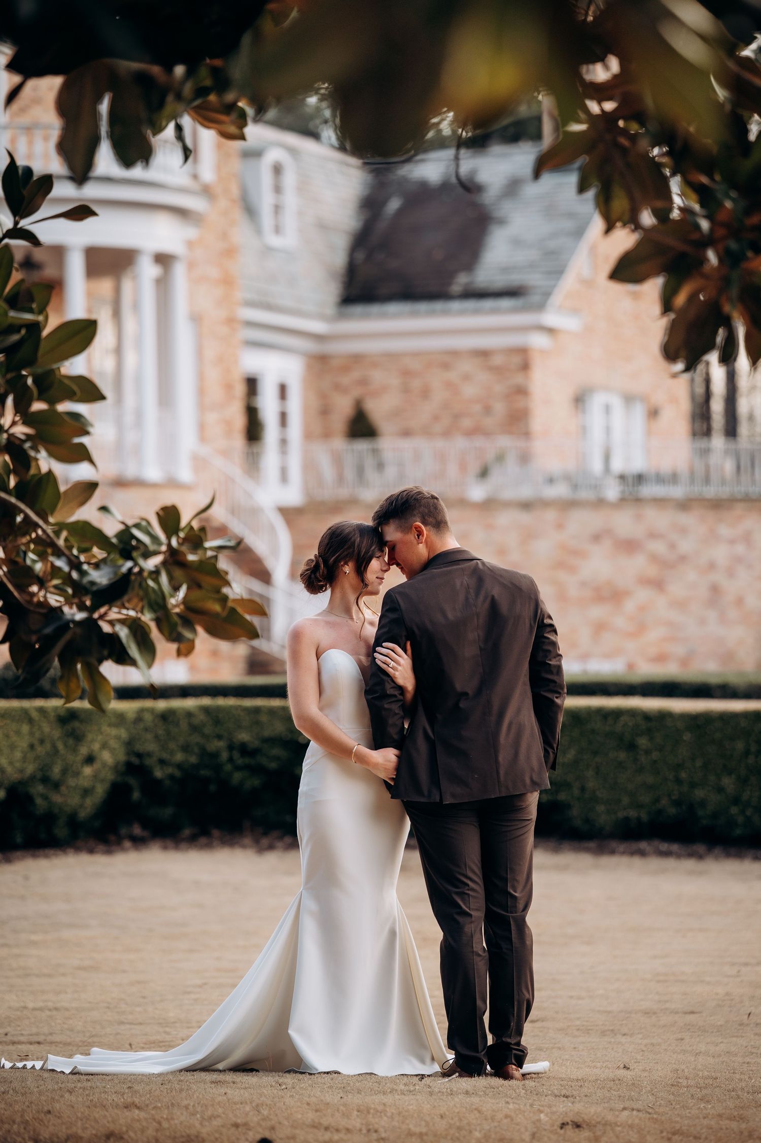 Romantic wedding couple sharing tender moment in garden with historic mansion backdrop.