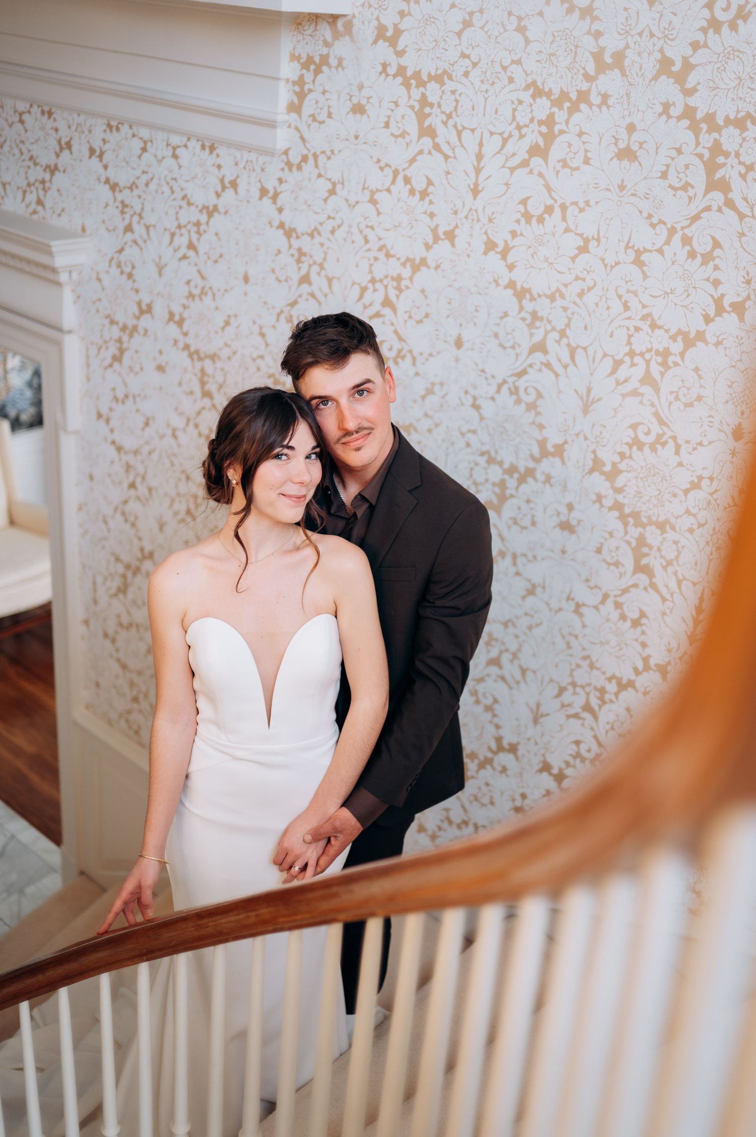 A romantic moment captured on an elegant staircase with floral wallpaper backdrop.
