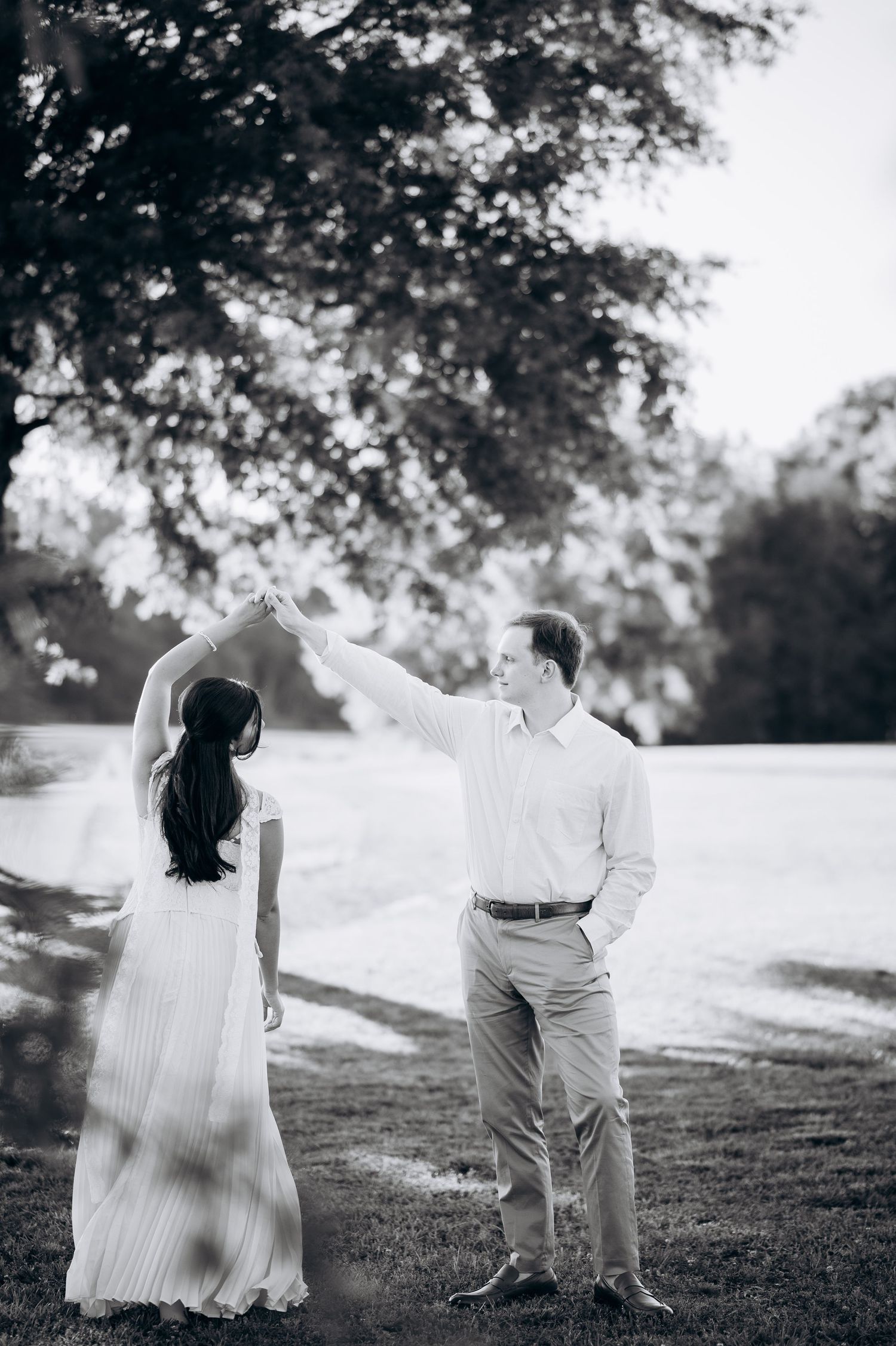 Black and white photo of a couple dancing outdoors, man spinning woman in white dress near a large tree.