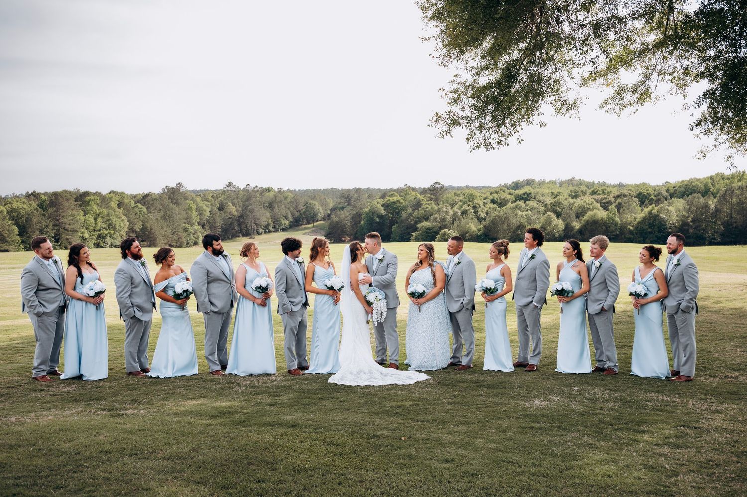 Wedding party in gray suits and light blue dresses standing on green lawn with trees in background.