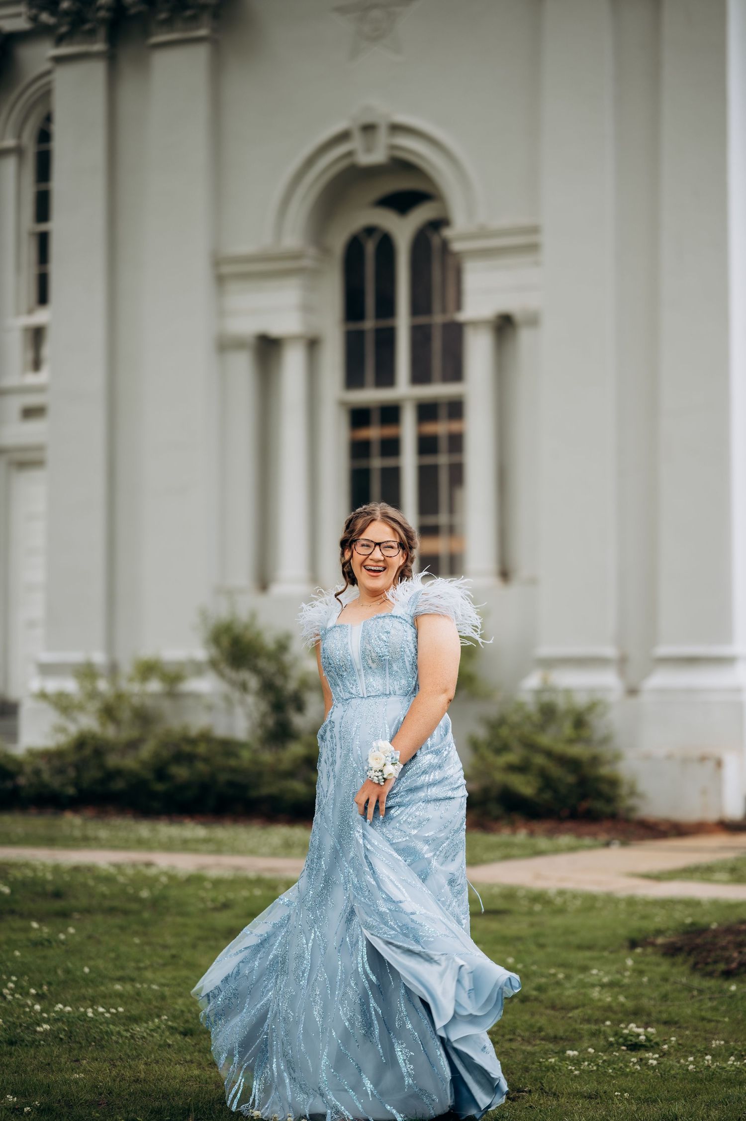 A young woman in a flowing light blue feathered prom gown laughs joyfully outdoors near a white building.