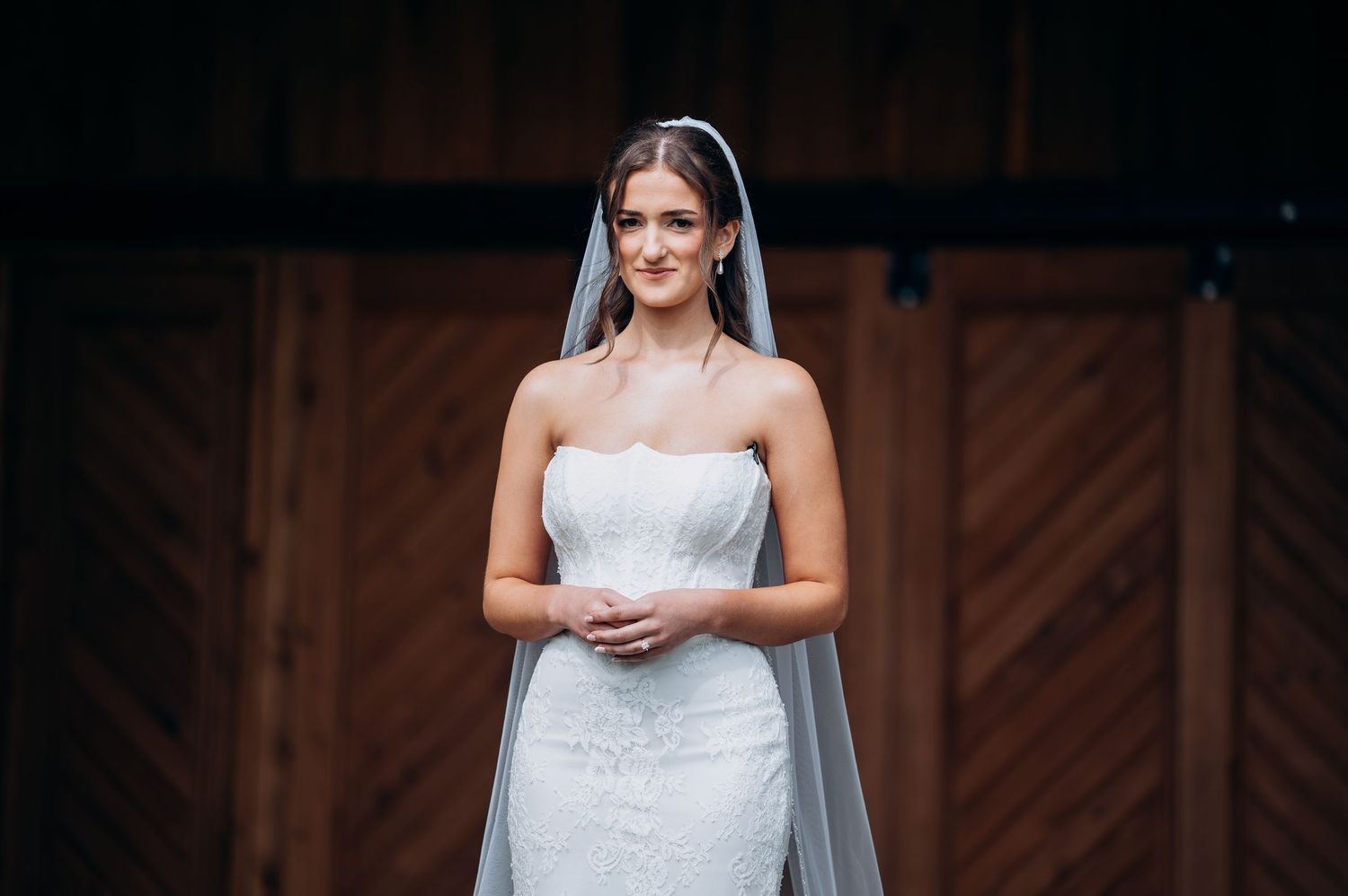 Beautiful bride in strapless lace mermaid gown with cathedral veil standing before rustic barn doors.