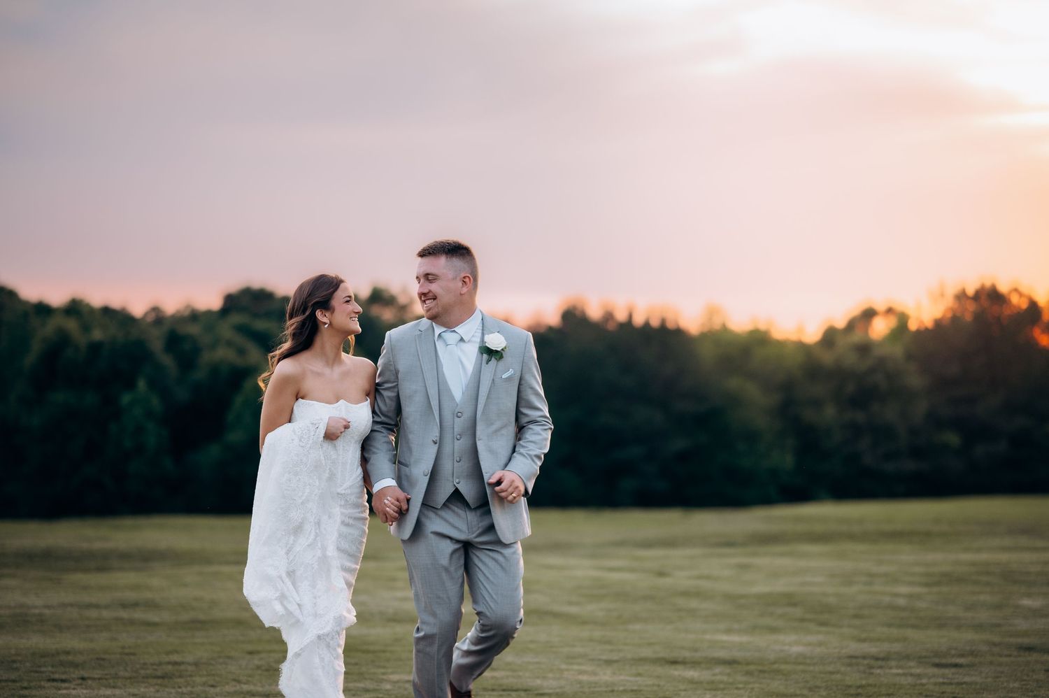 Bride and groom walking hand in hand at sunset in a lush green field, wearing white lace gown and gray suit.