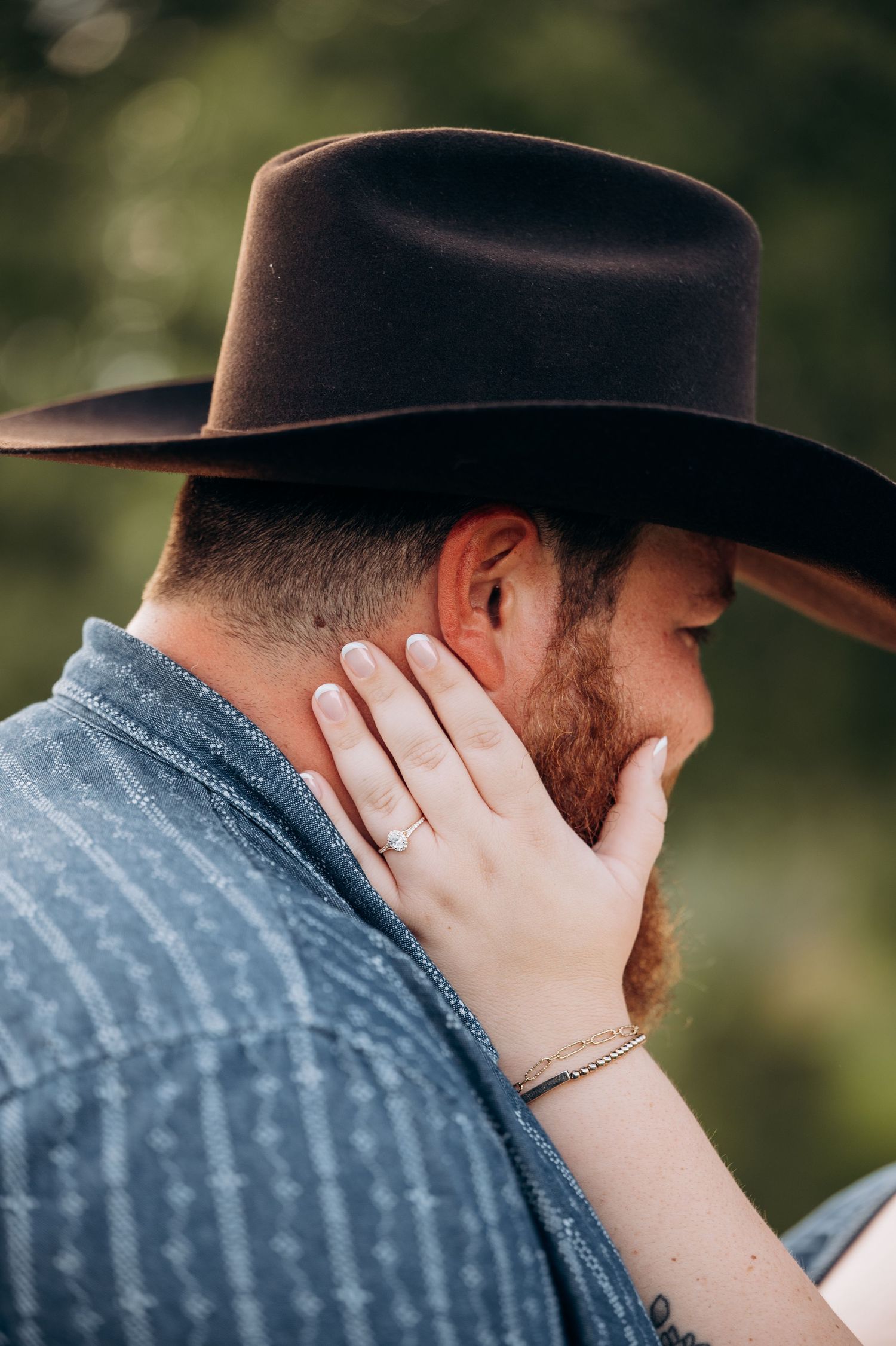 Close-up of woman's hand with engagement ring resting on bearded man's face wearing a brown cowboy hat.