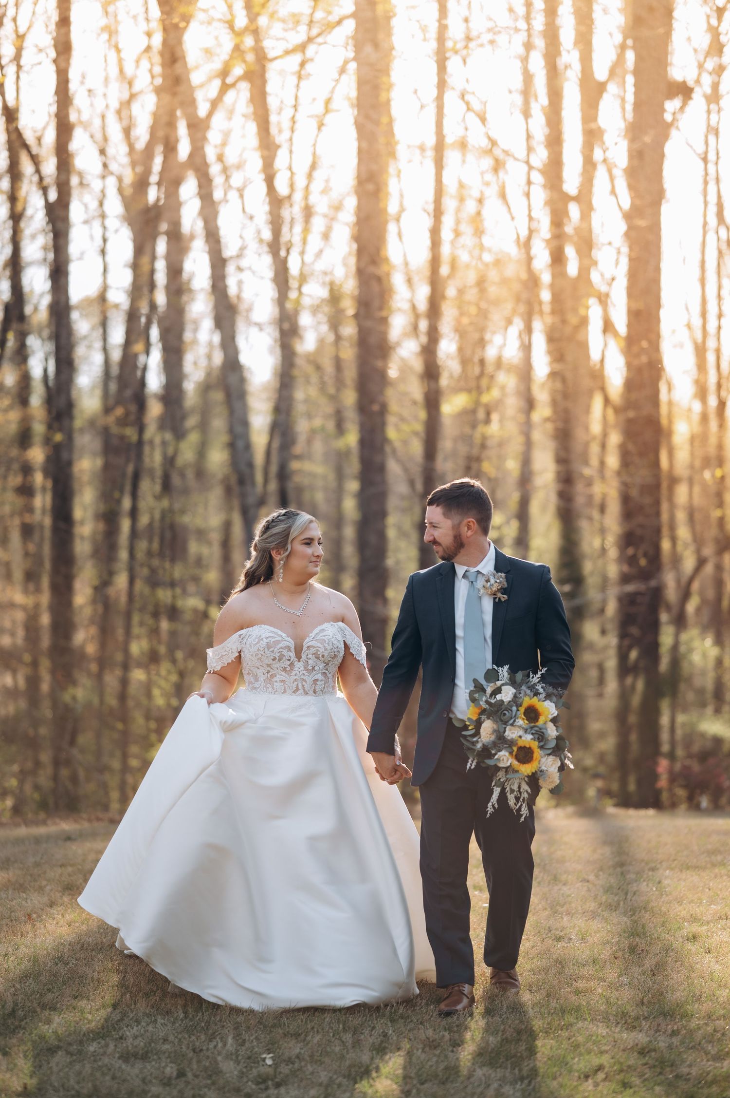 Newlyweds walk hand in hand through a sunlit forest during golden hour.