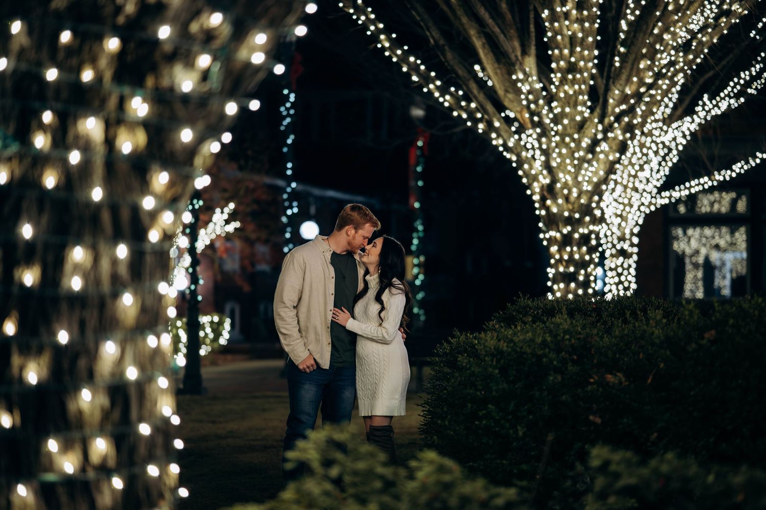 Romantic couple sharing a moment surrounded by twinkling fairy lights on trees at night.