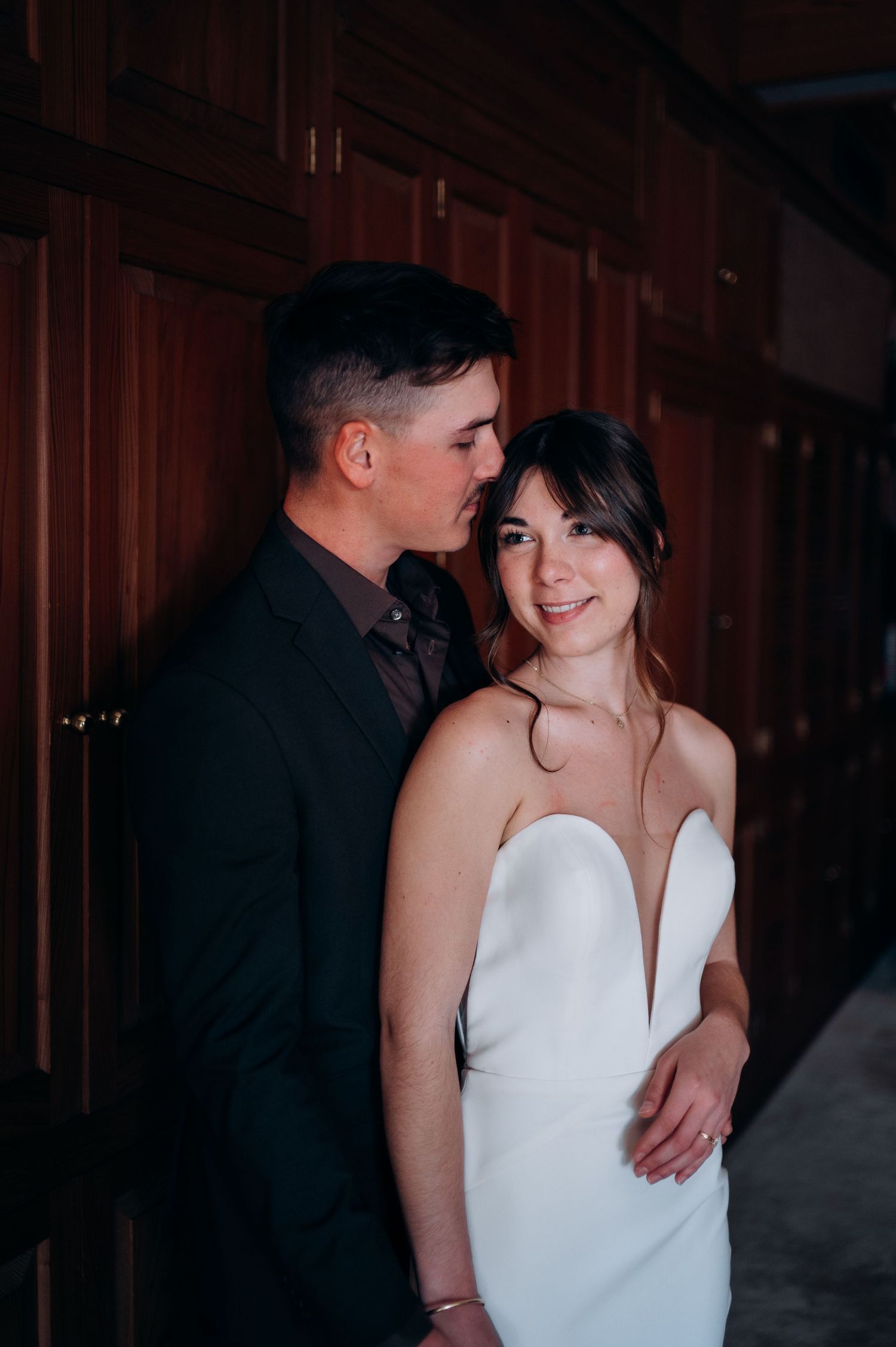 Groom whispering to smiling bride in white strapless gown against dark wood paneling.