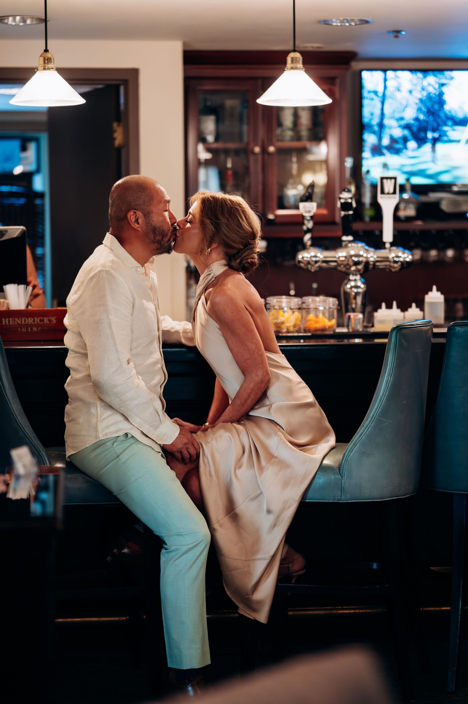 Couple sharing a romantic kiss while seated at an elegant bar with pendant lights and teal chairs.