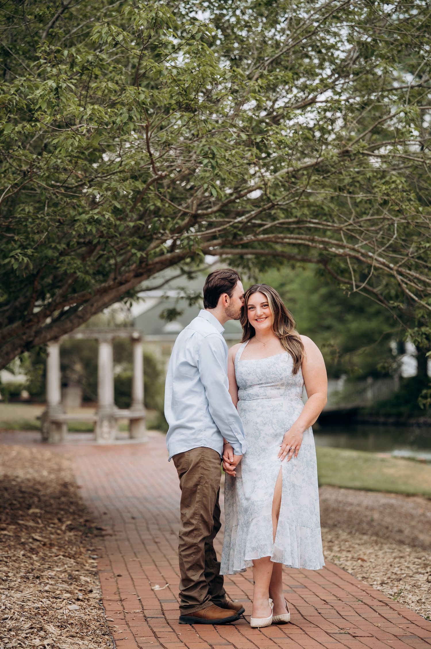 Couple holding hands on a brick path beneath a sprawling tree near a white gazebo.