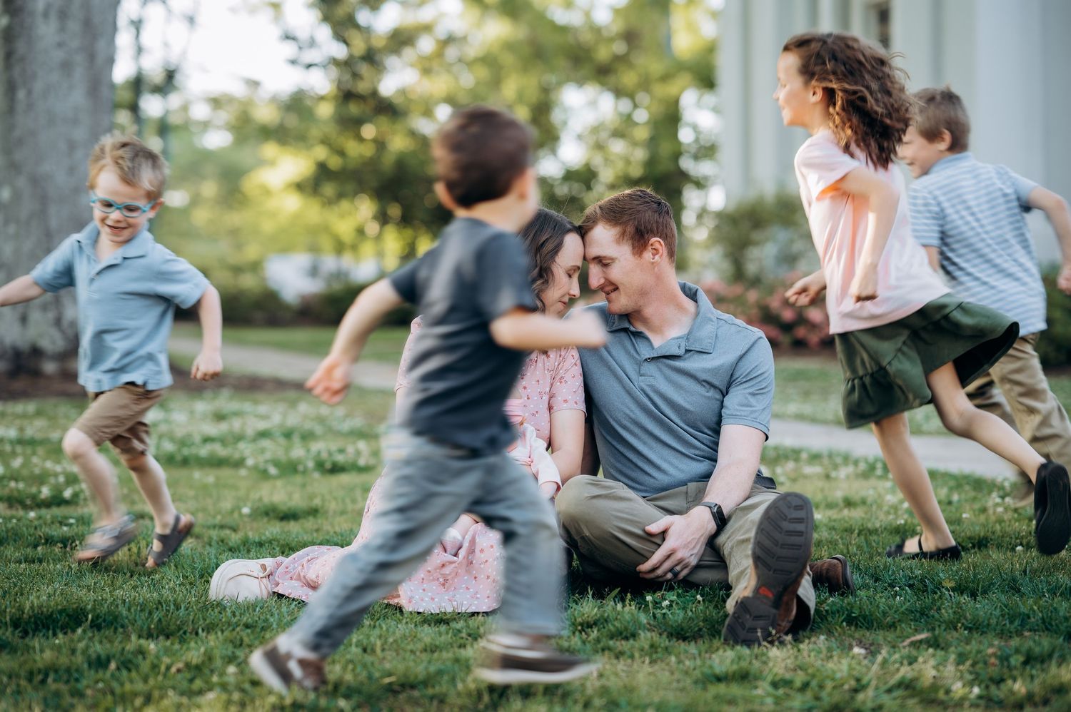 A family sits on green grass while children run and play around their smiling parents outdoors.