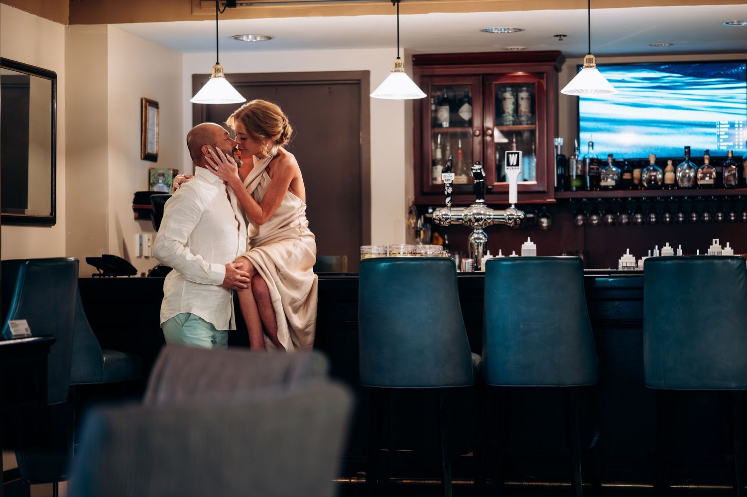 Couple sharing a tender kiss at a sophisticated bar with blue velvet stools.